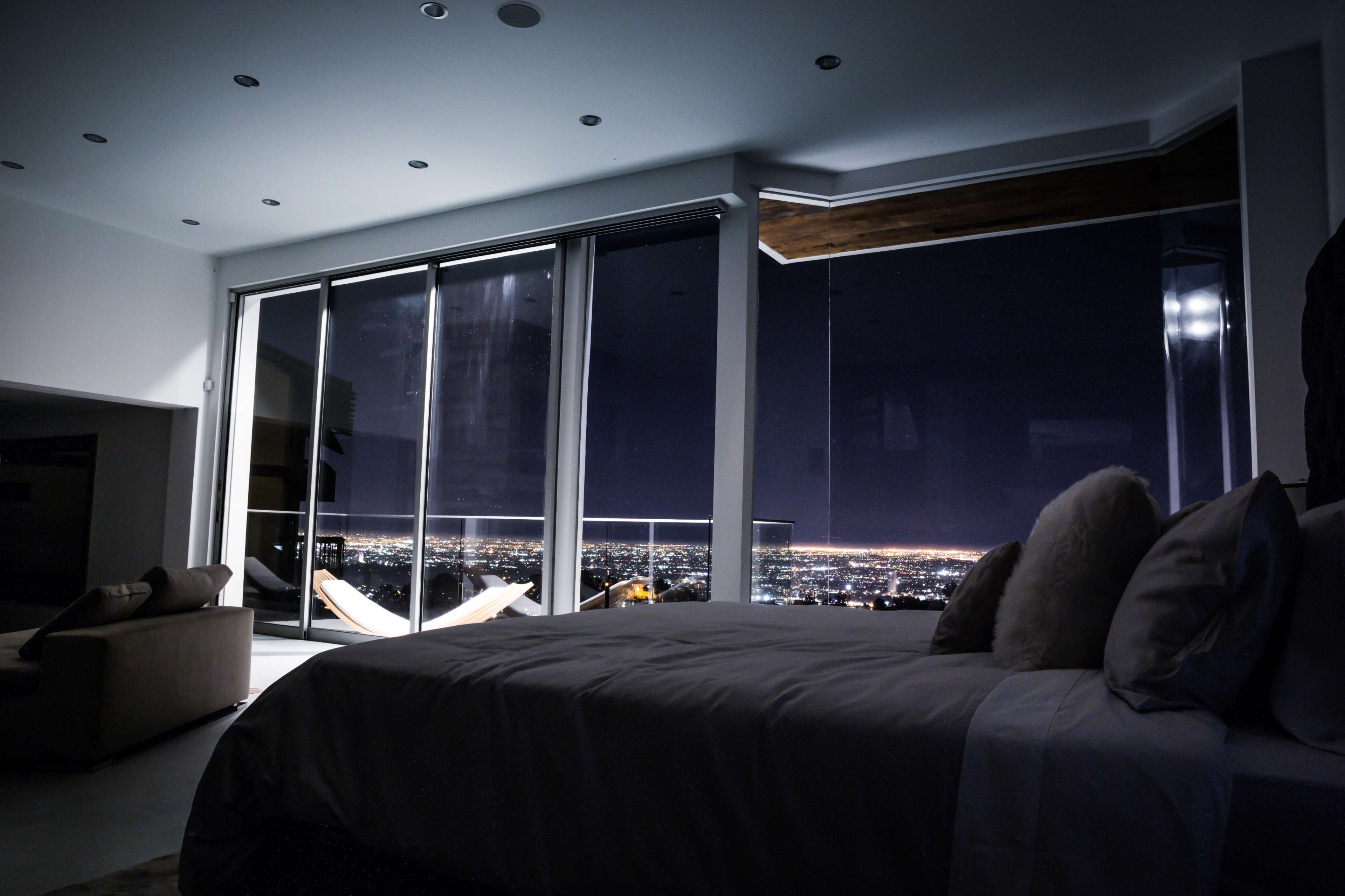 A modern bedroom features a large window framing a panoramic view of a city skyline illuminated at night.