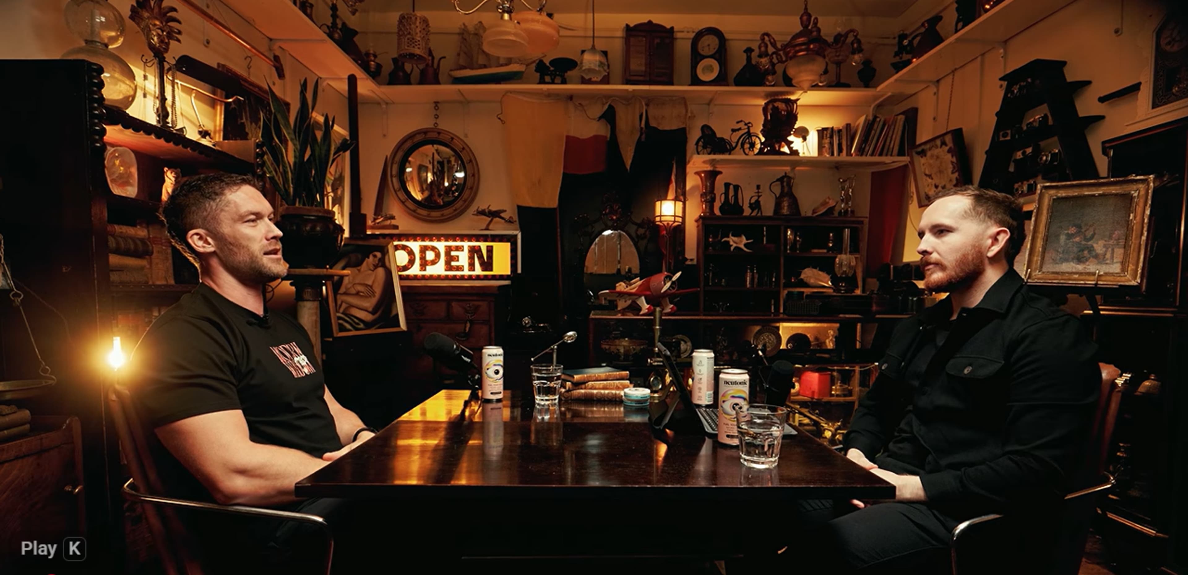 Two men sit at a table in a cluttered room decorated with various antiques and a sign that reads "OPEN."