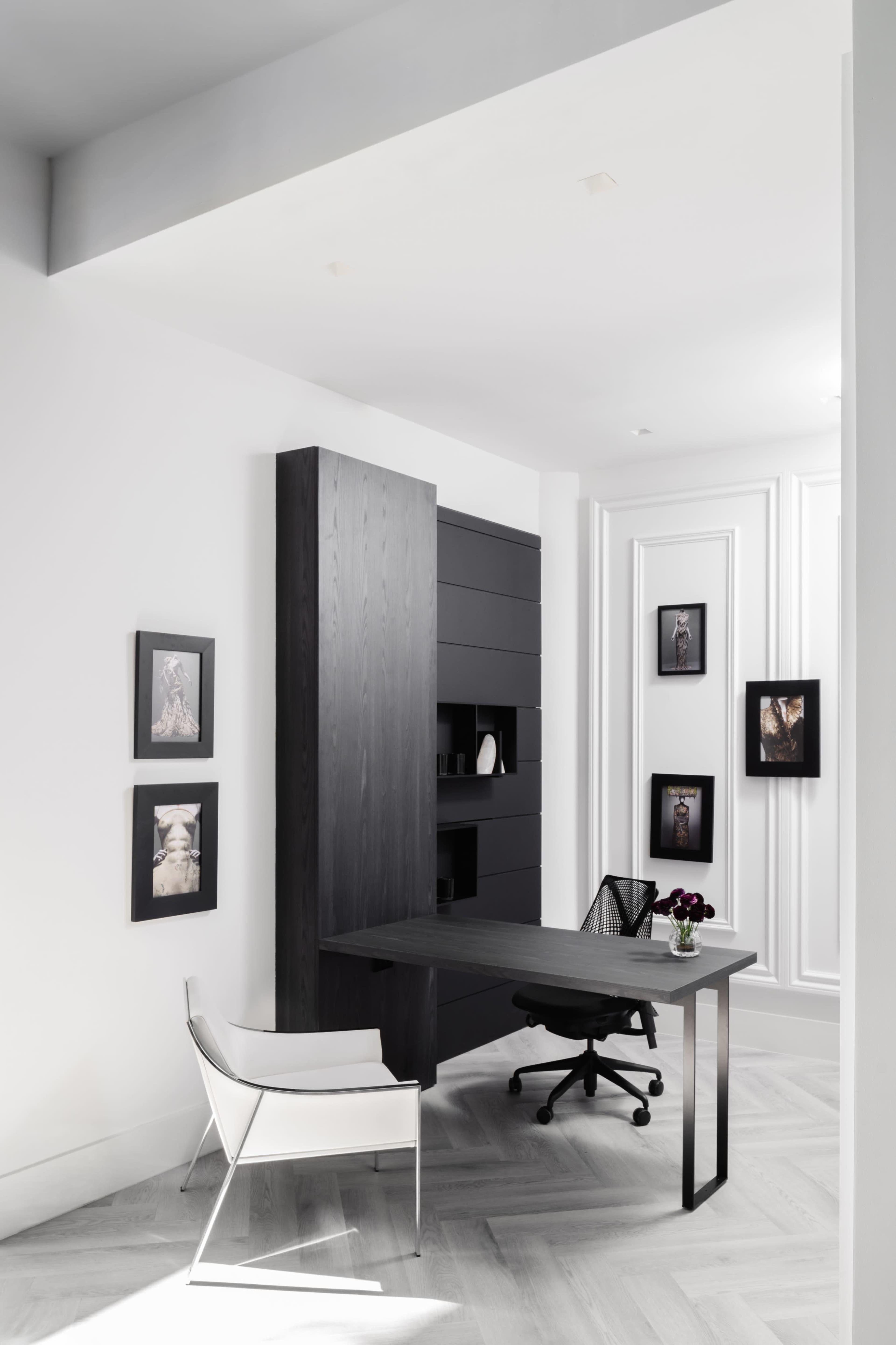 A minimalist office space features a black desk and chair, surrounded by white walls adorned with framed photographs.