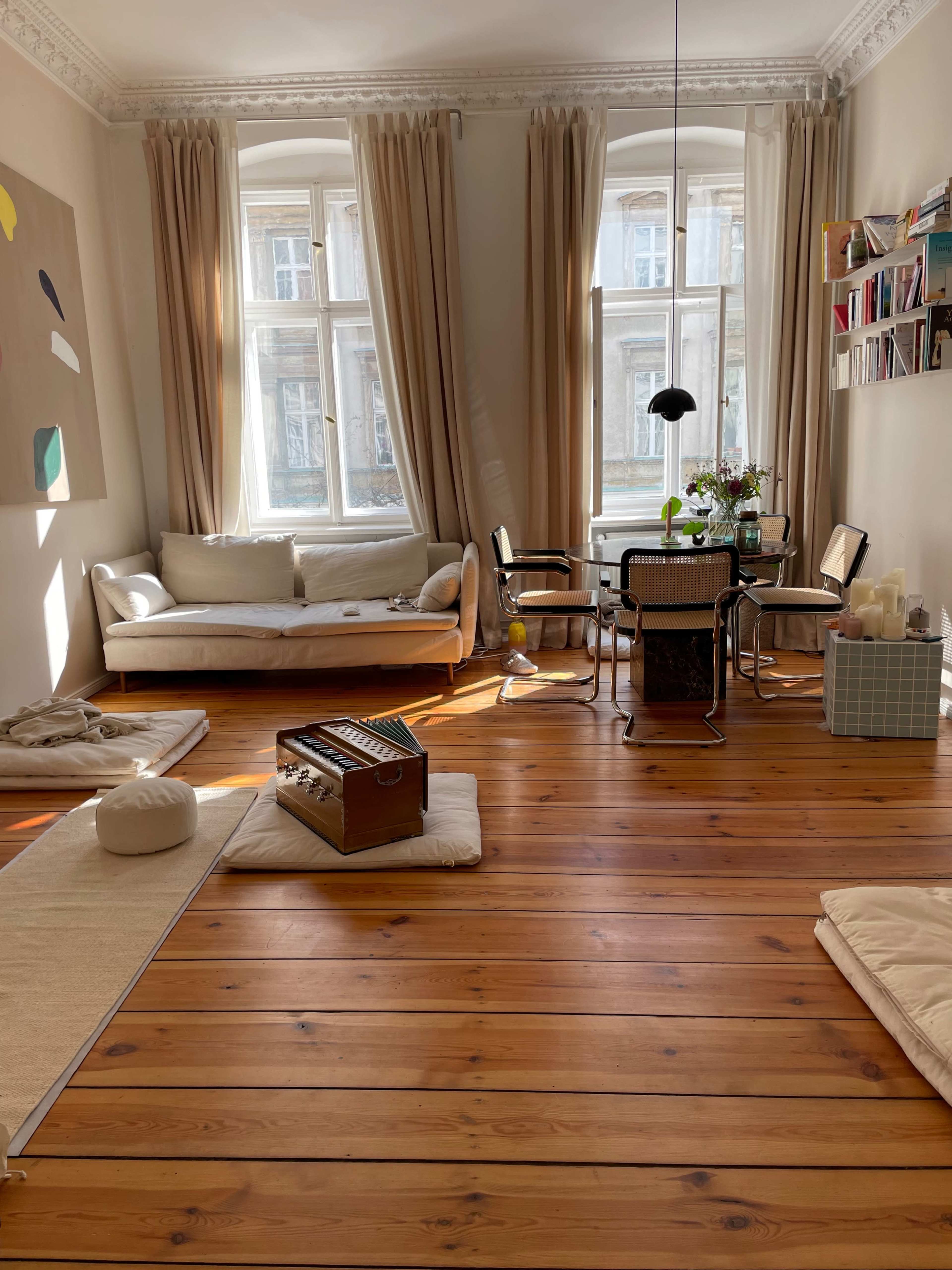 The room features a light-colored sofa, a dining table with chairs, and wooden flooring, illuminated by natural light coming through the large windows.