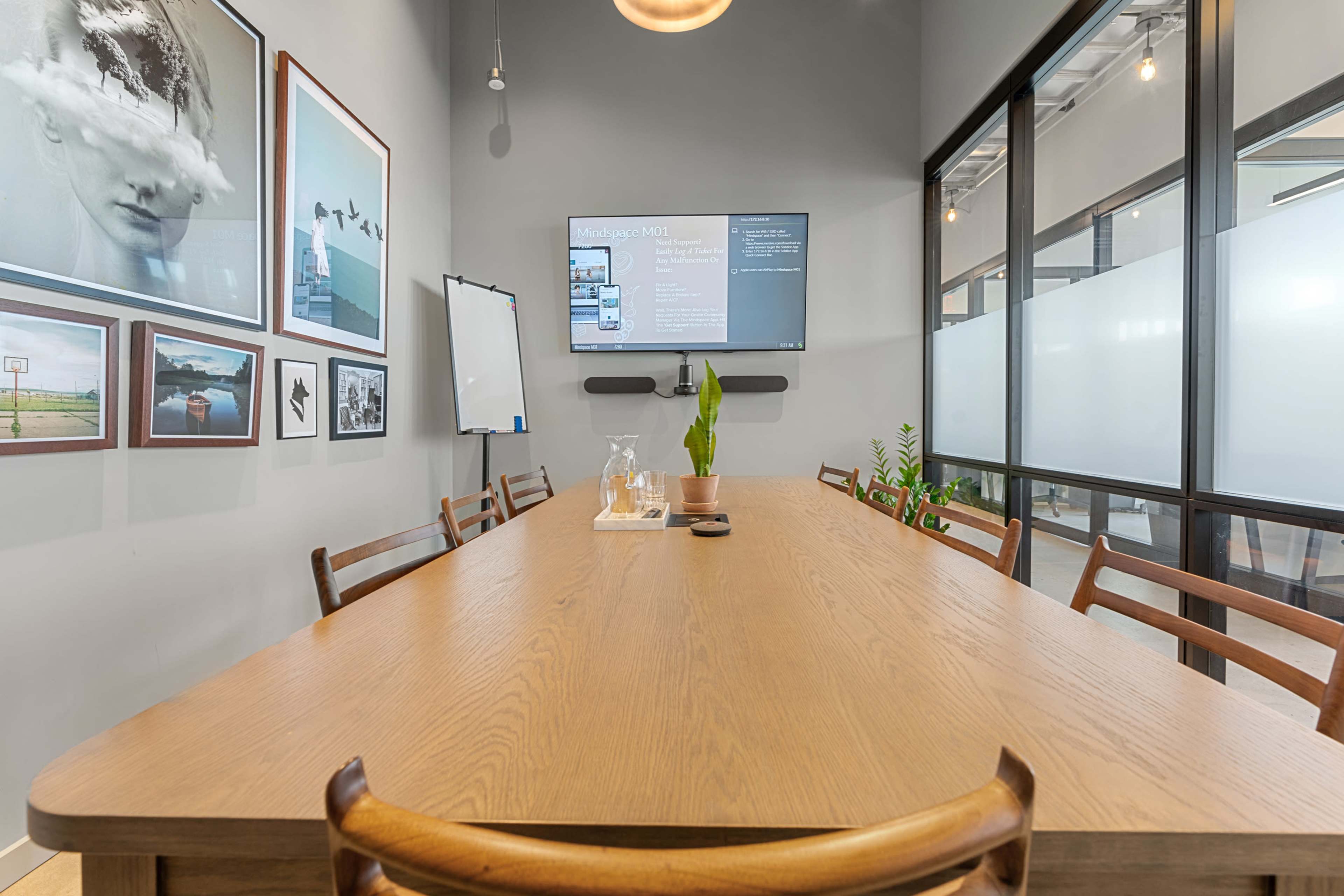 A modern conference room features a long wooden table, framed photographs on the walls, a digital screen displaying information, and greenery in pots.