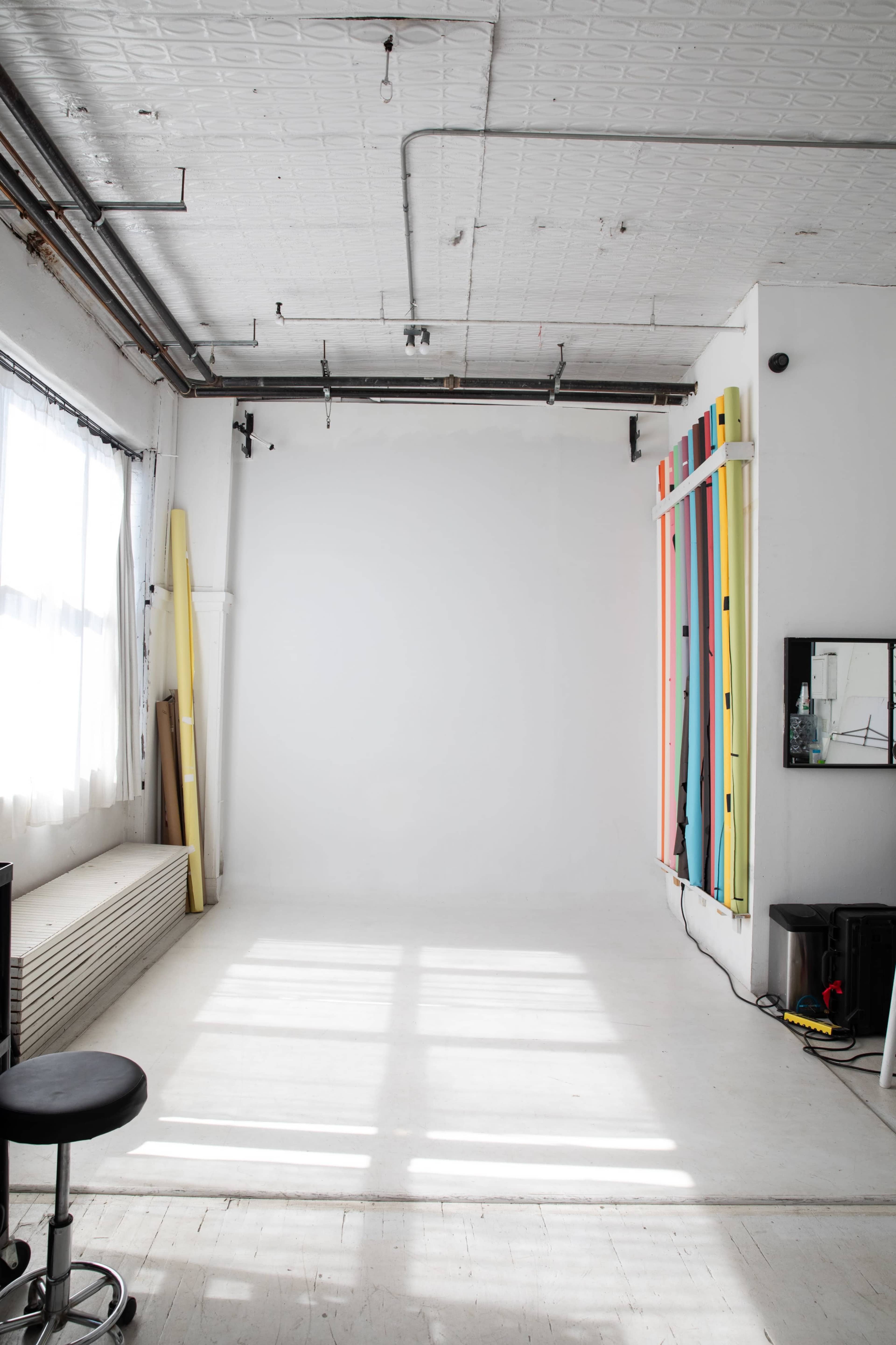 The image shows a spacious, minimalist studio with a white wall, natural light coming from a window, and colorful vertical poles arranged on one side.