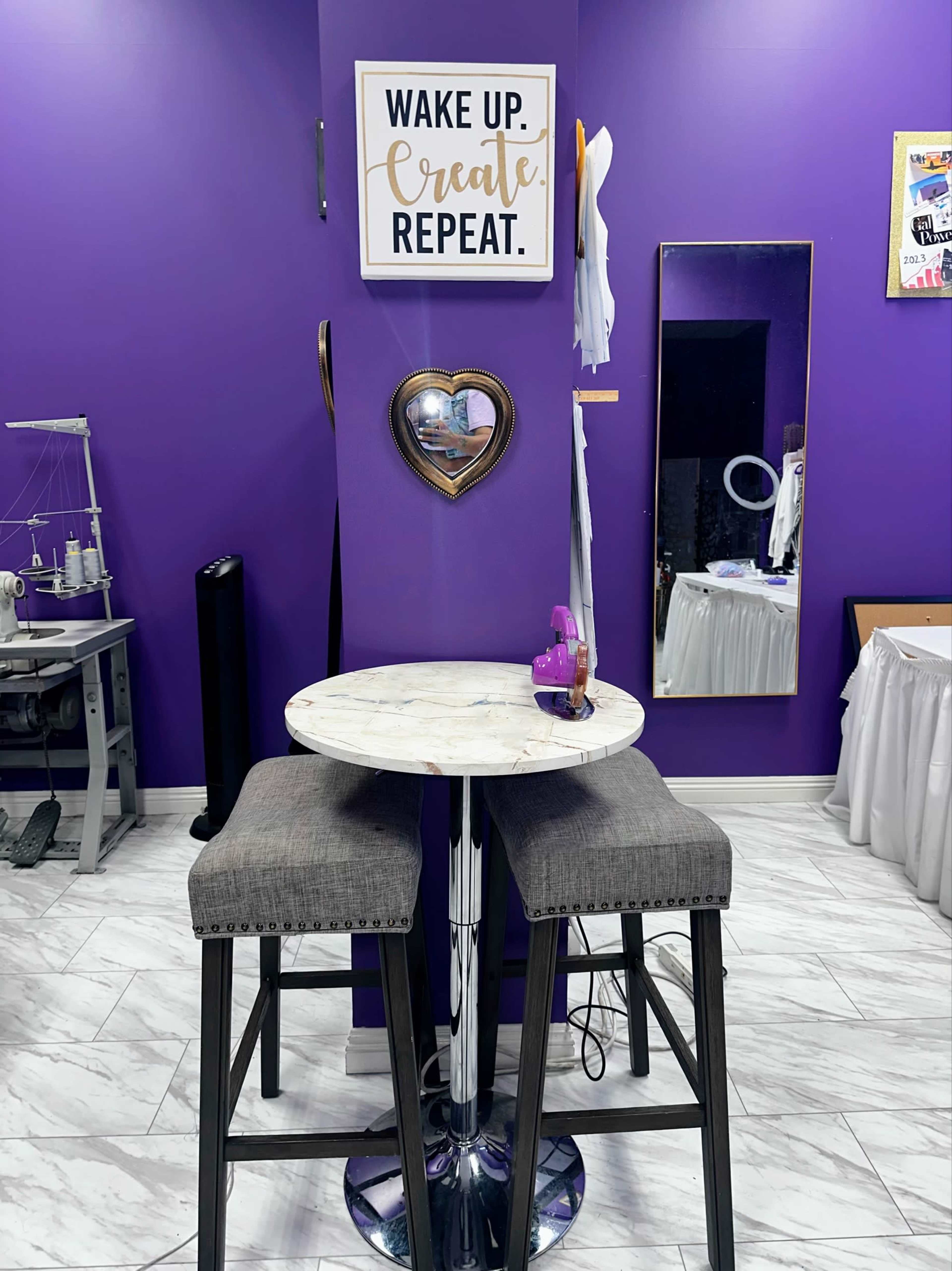 A small round table with two stools is positioned against a purple wall adorned with a motivational sign and a heart-shaped mirror.