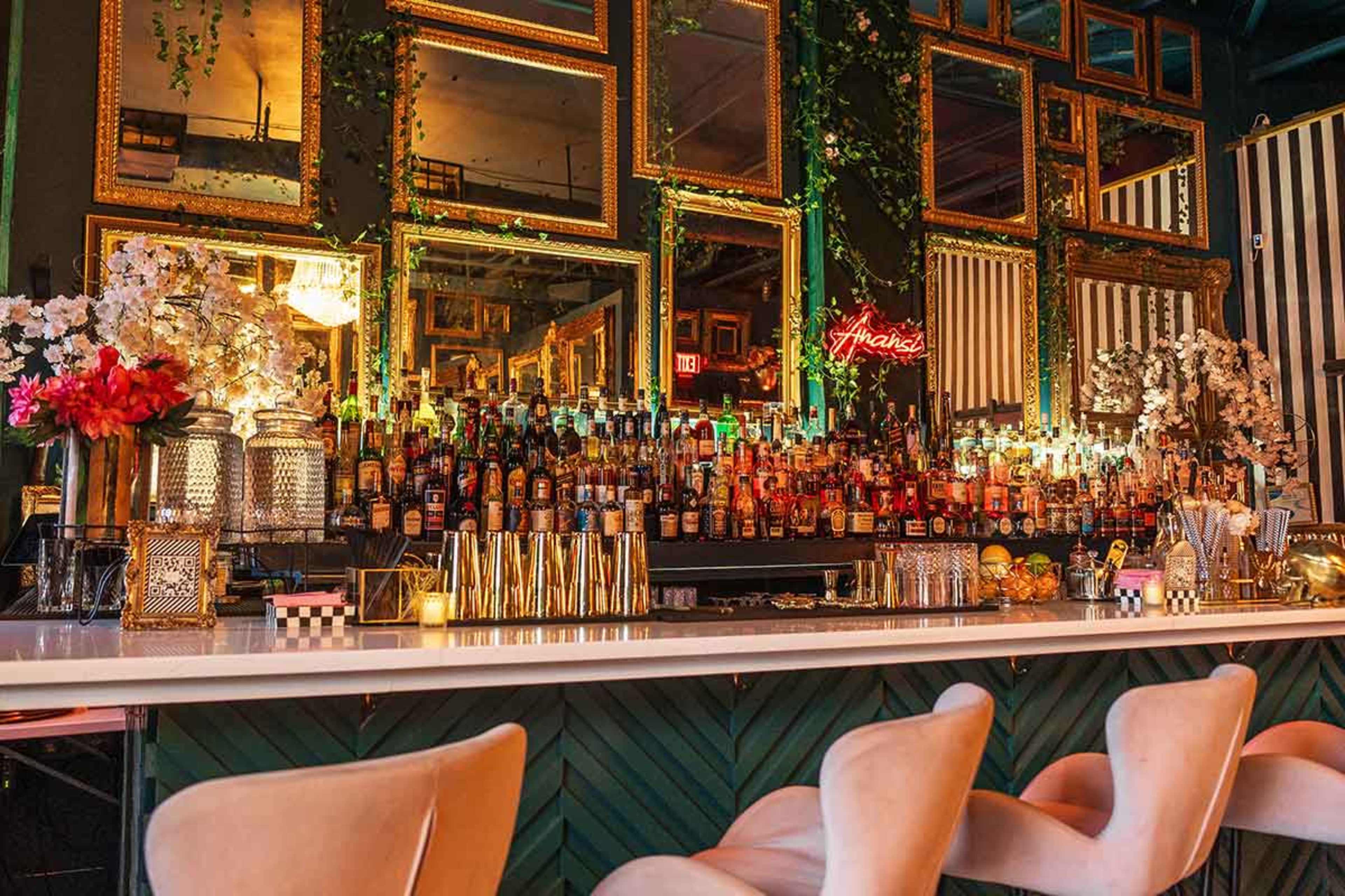The image shows a stylish bar area featuring a long white countertop, an array of colorful bottles organized on shelves behind it, and multiple ornate golden mirrors reflecting the scene.