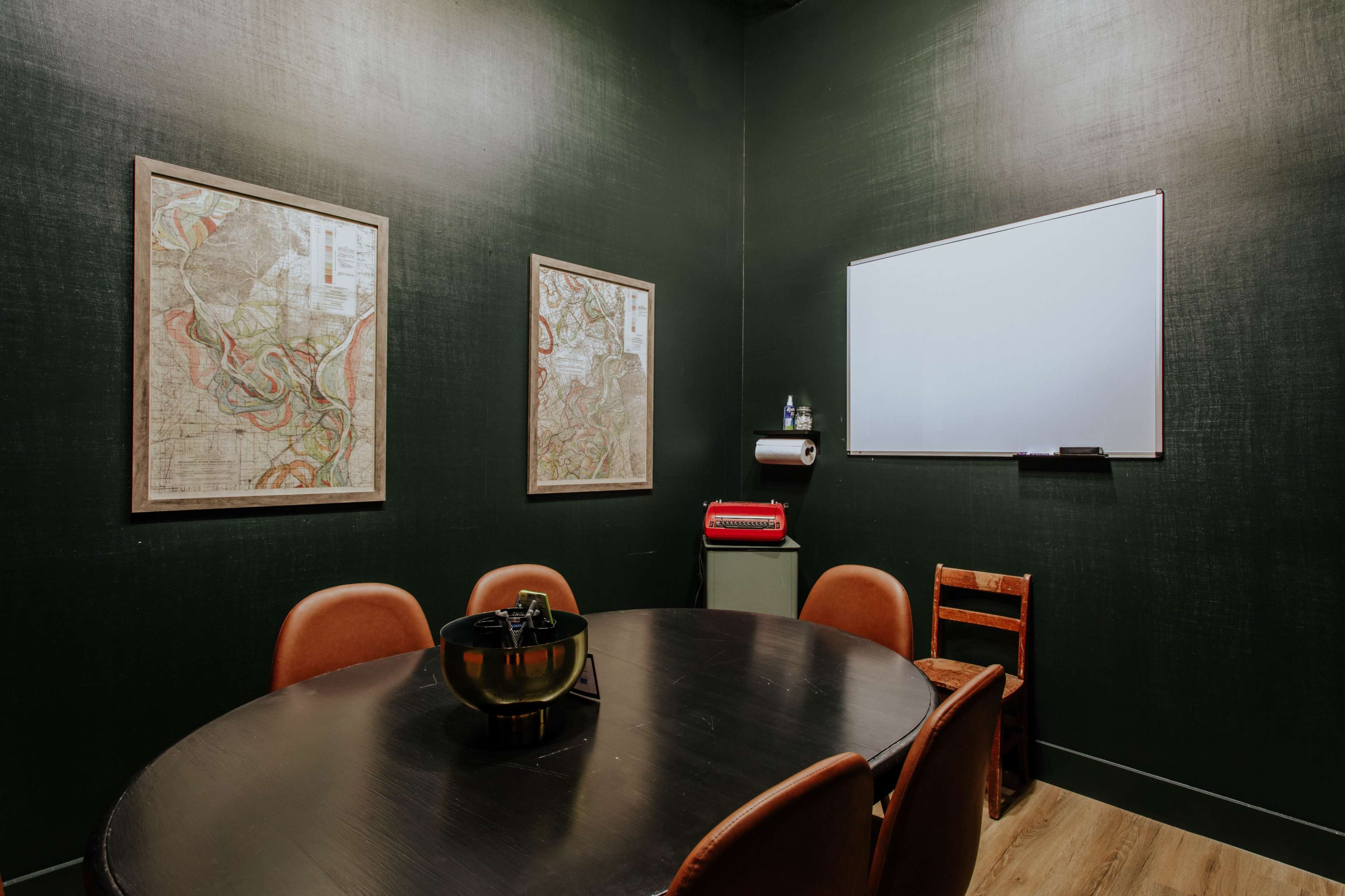 A small, dark green meeting room with a round table, brown chairs, framed maps on the walls, and a whiteboard.
