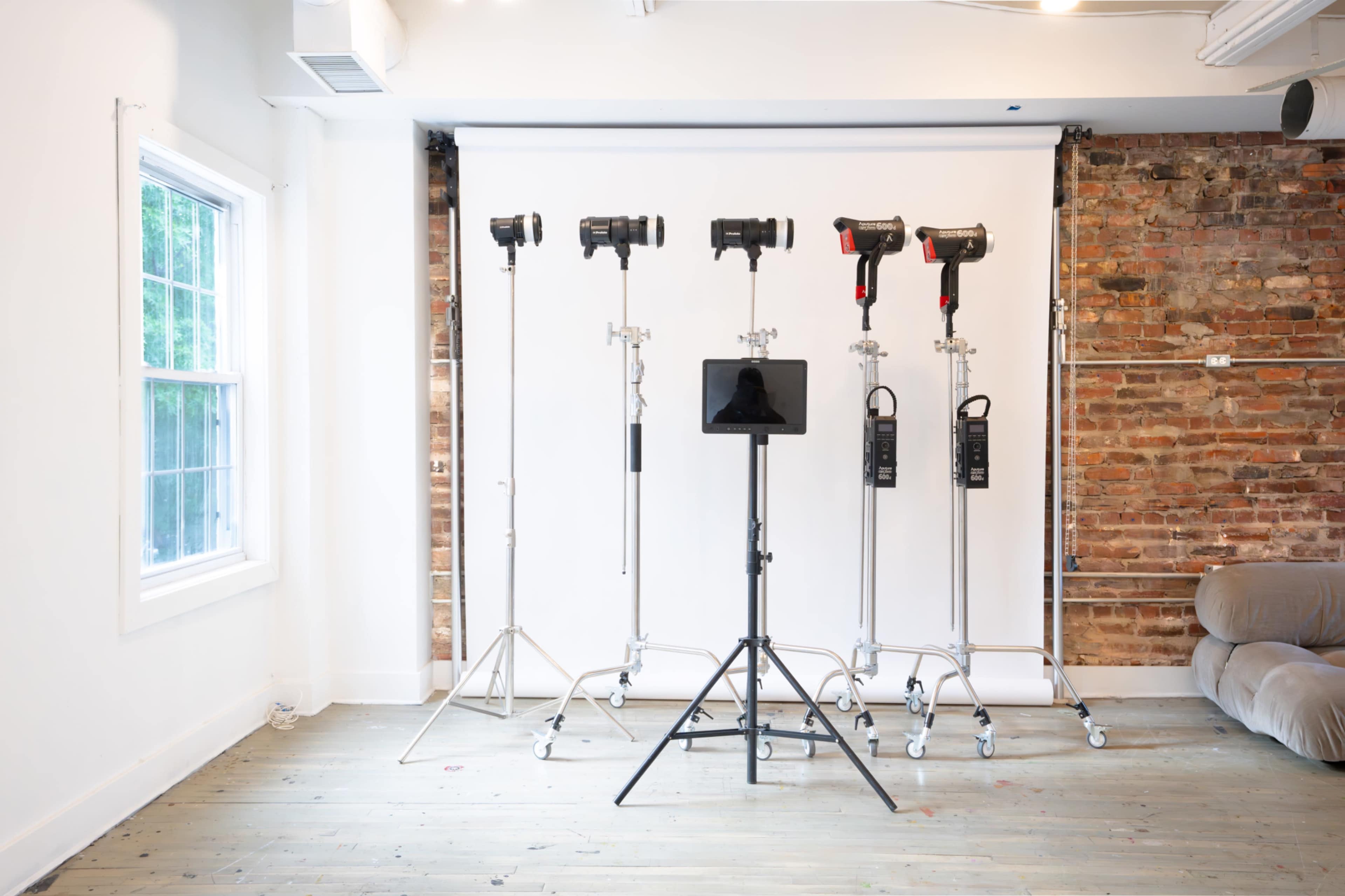 The image shows a photography studio setup featuring multiple light stands, a backdrop, and a stand for equipment in a room with wooden flooring and brick walls.