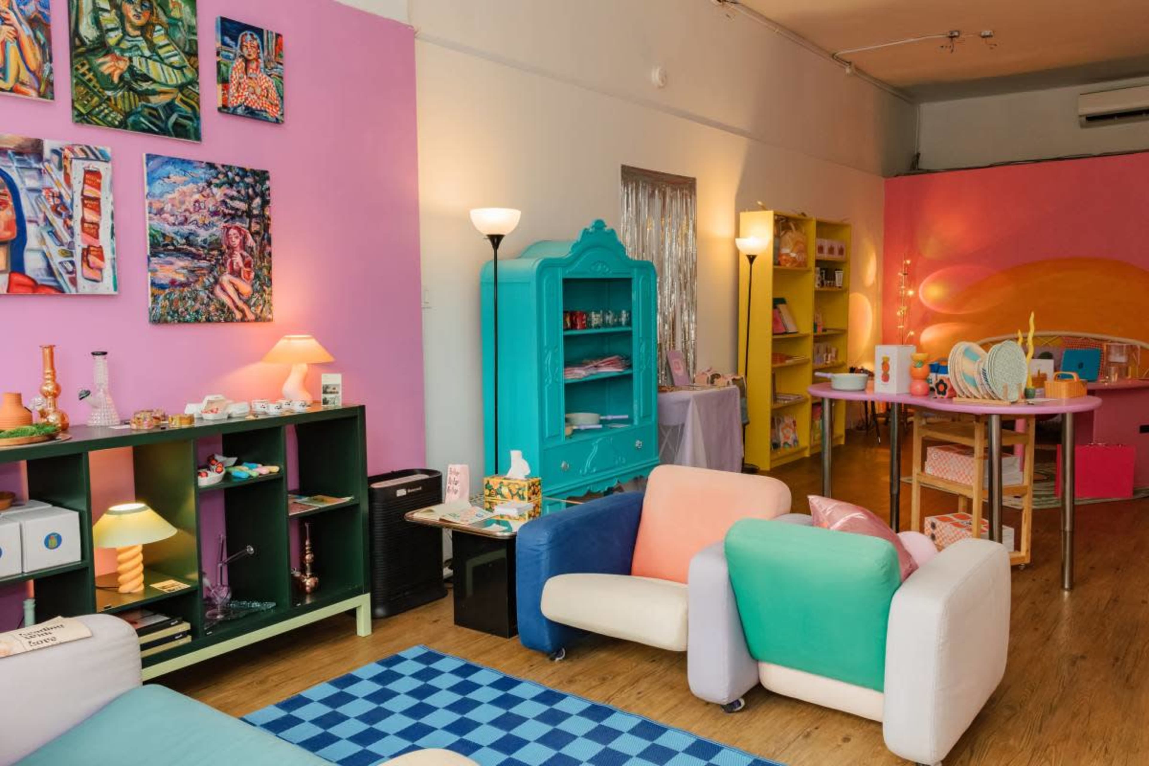 The image shows a colorful room decorated with vibrant wall paintings, playful furniture in pastel colors, and various shelves displaying products.