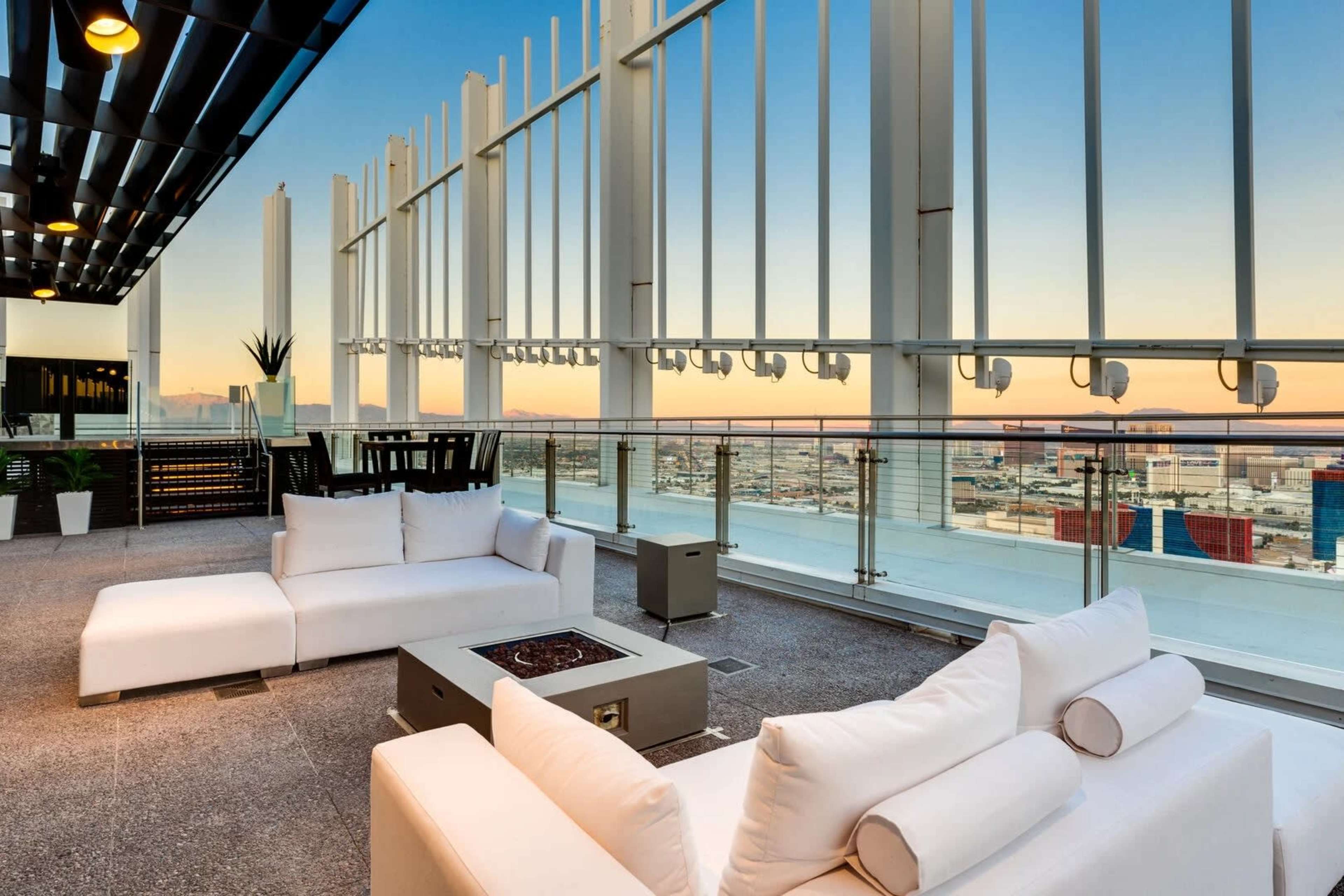 The image shows a modern rooftop lounge area with white furniture, a fire pit, and a view of the city skyline at sunset.