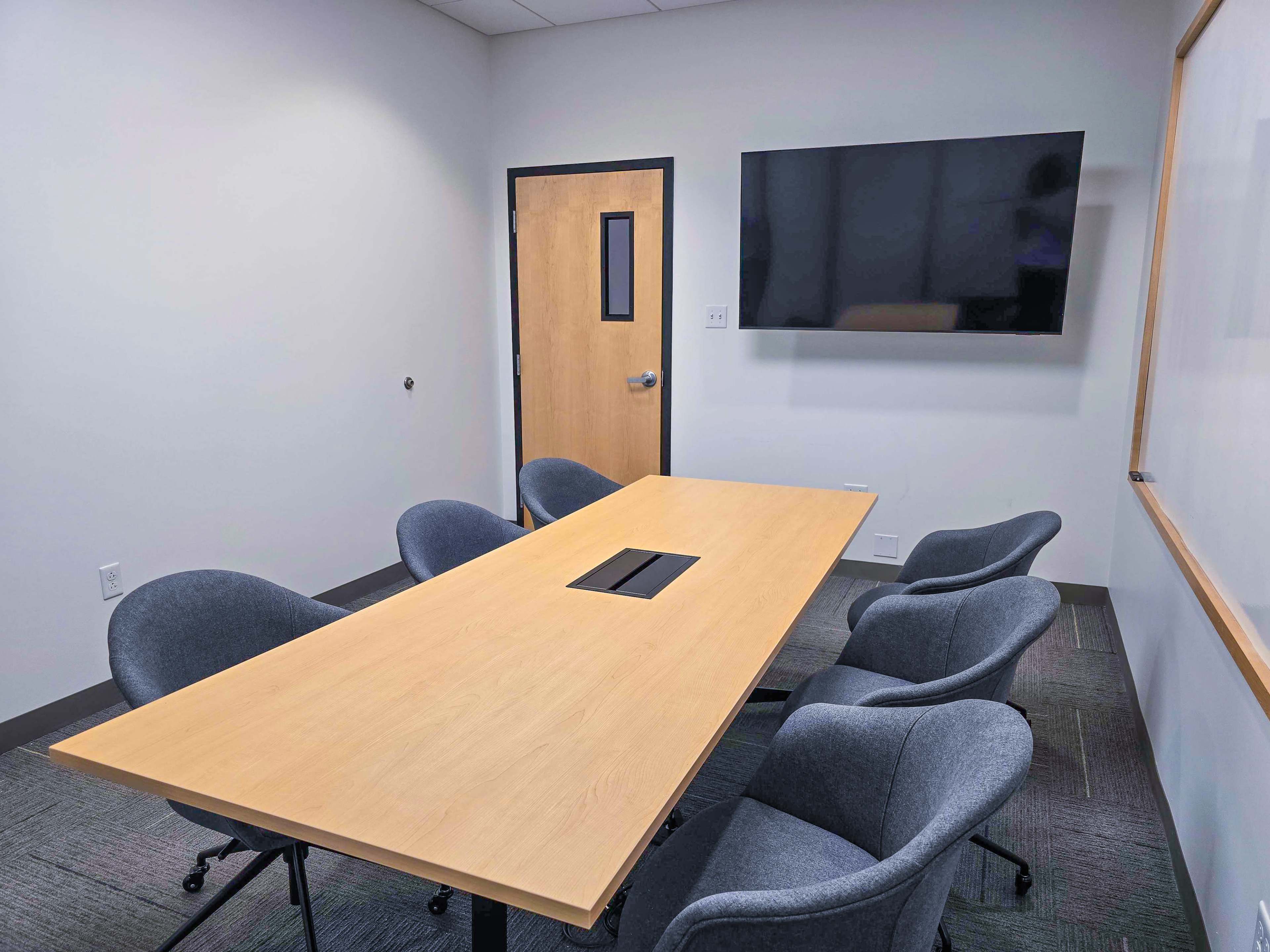 The image shows a modern conference room with a rectangular wooden table, six gray chairs, a large television mounted on the wall, and a whiteboard.