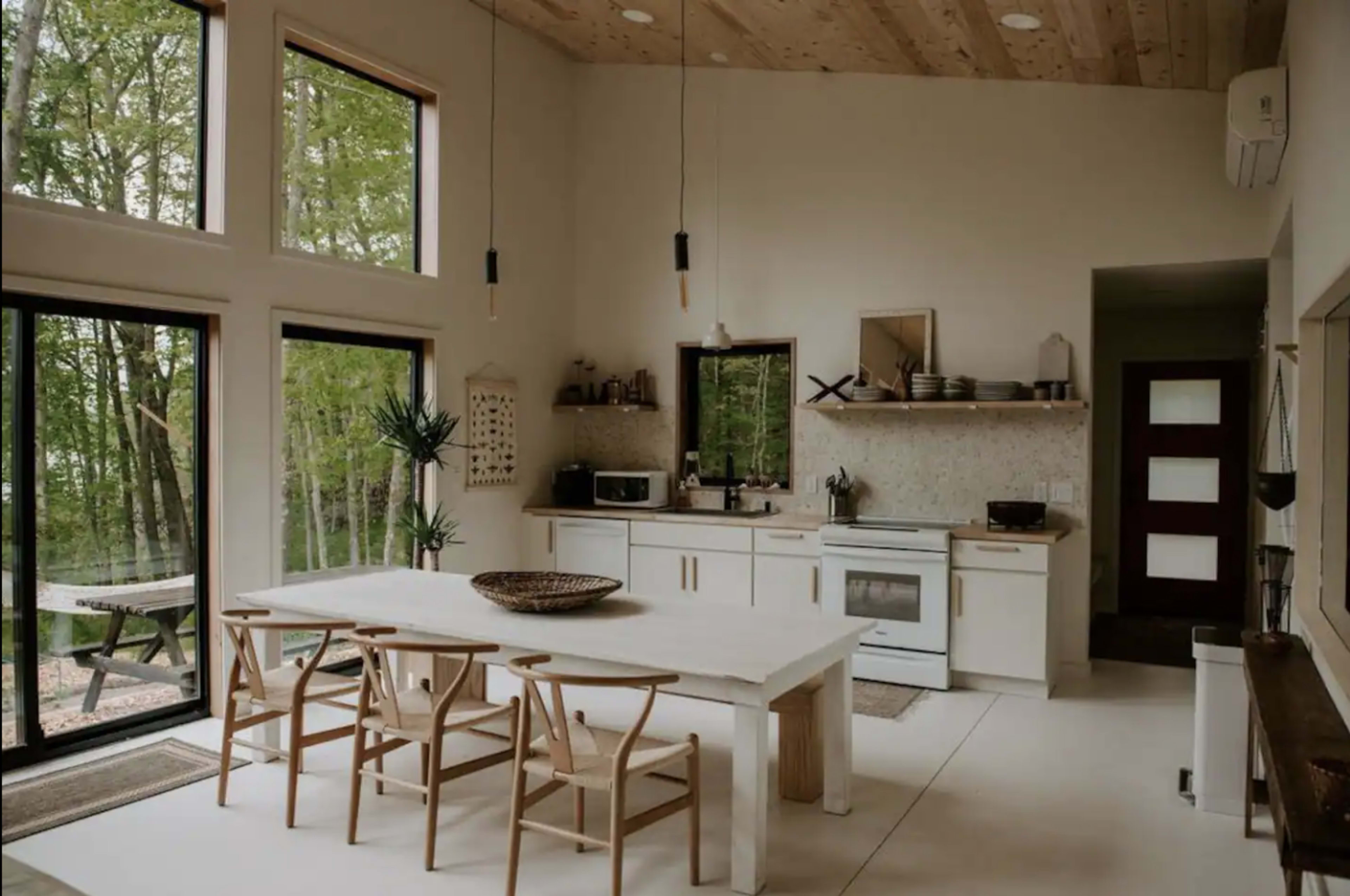 A modern kitchen features a large dining table with wooden chairs, large windows overlooking a forest, and minimalist appliances.