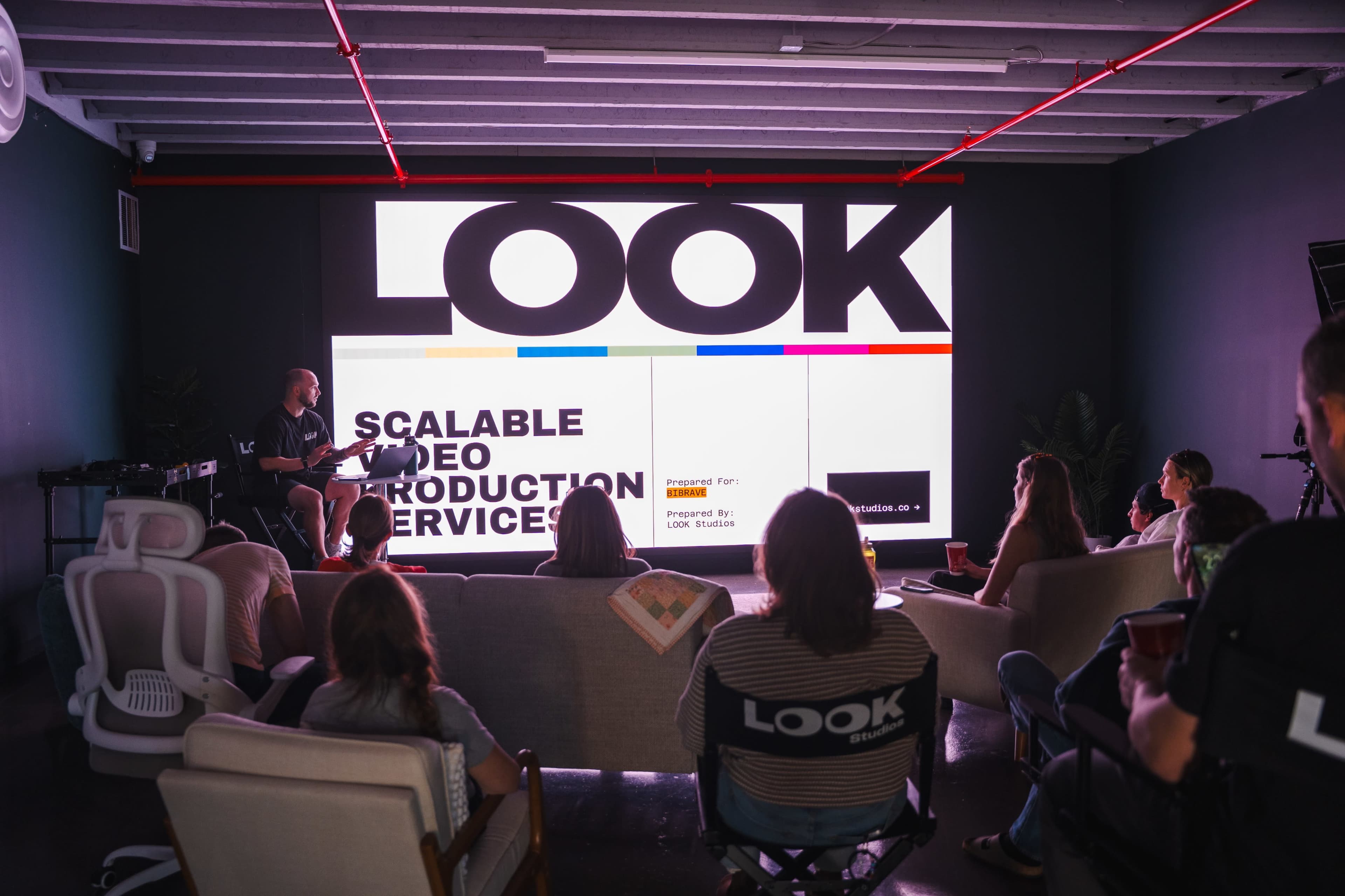 A group of people is seated in a modern meeting space, facing a large screen that displays the word "LOOK" and the phrase "SCALABLE VIDEO PRODUCTION SERVICES."