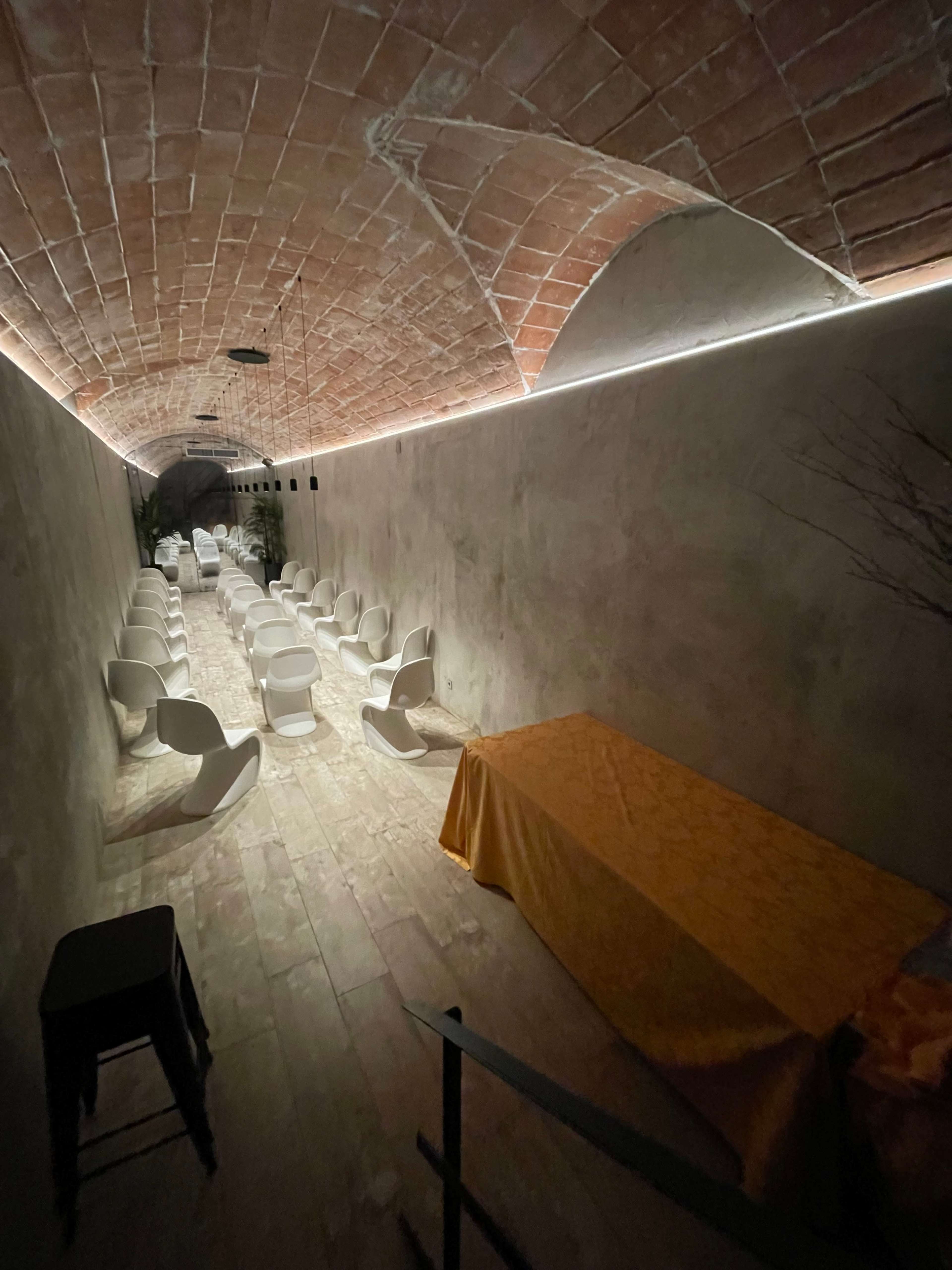 A long, narrow underground space with a series of white chairs arranged facing a table covered with a yellow tablecloth.