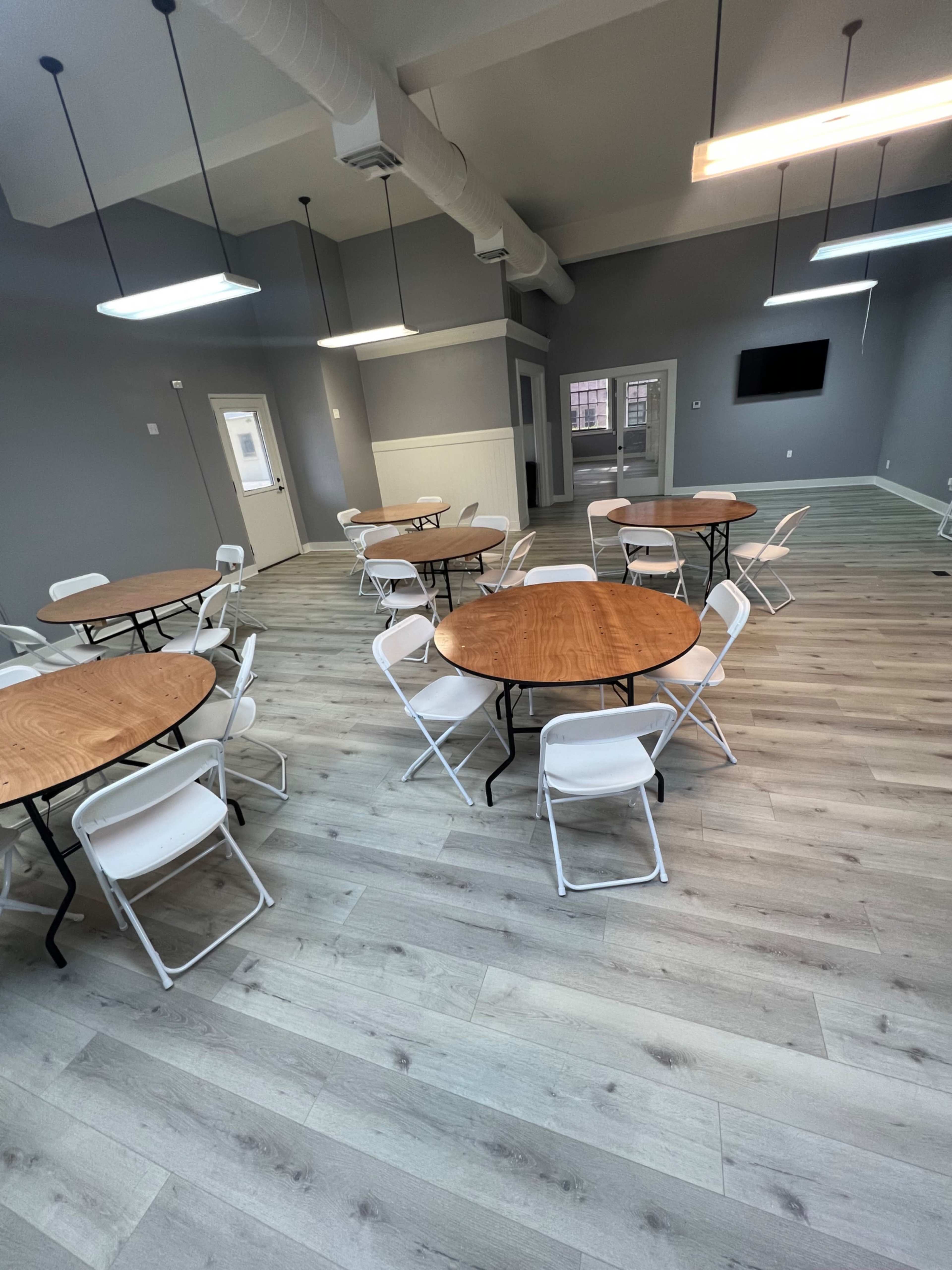 The image shows a spacious room with multiple round wooden tables and white folding chairs arranged on a light-colored wooden floor.
