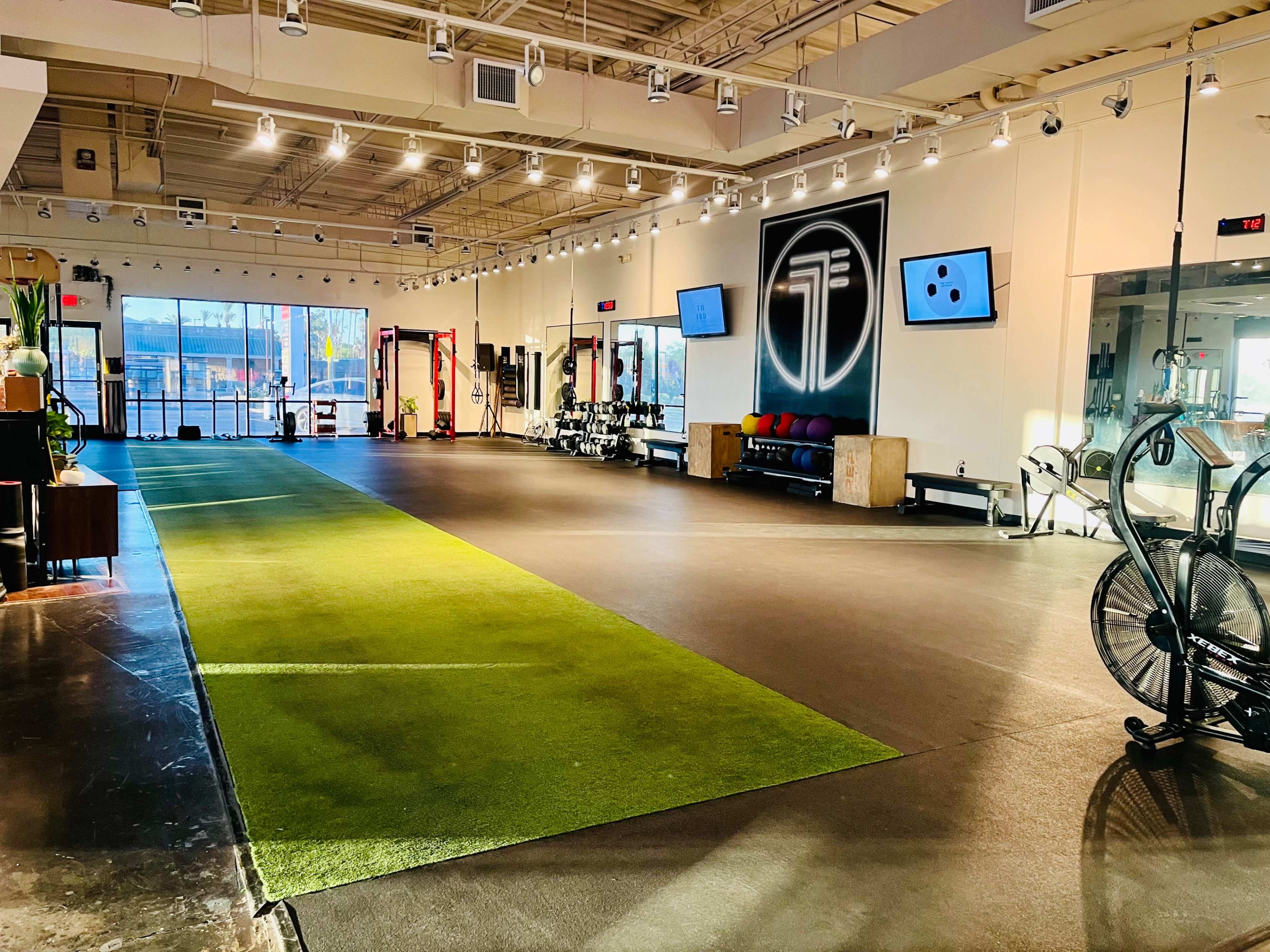 A spacious, well-lit gym with green turf in the center, exercise equipment along the walls, and large windows allowing natural light in.