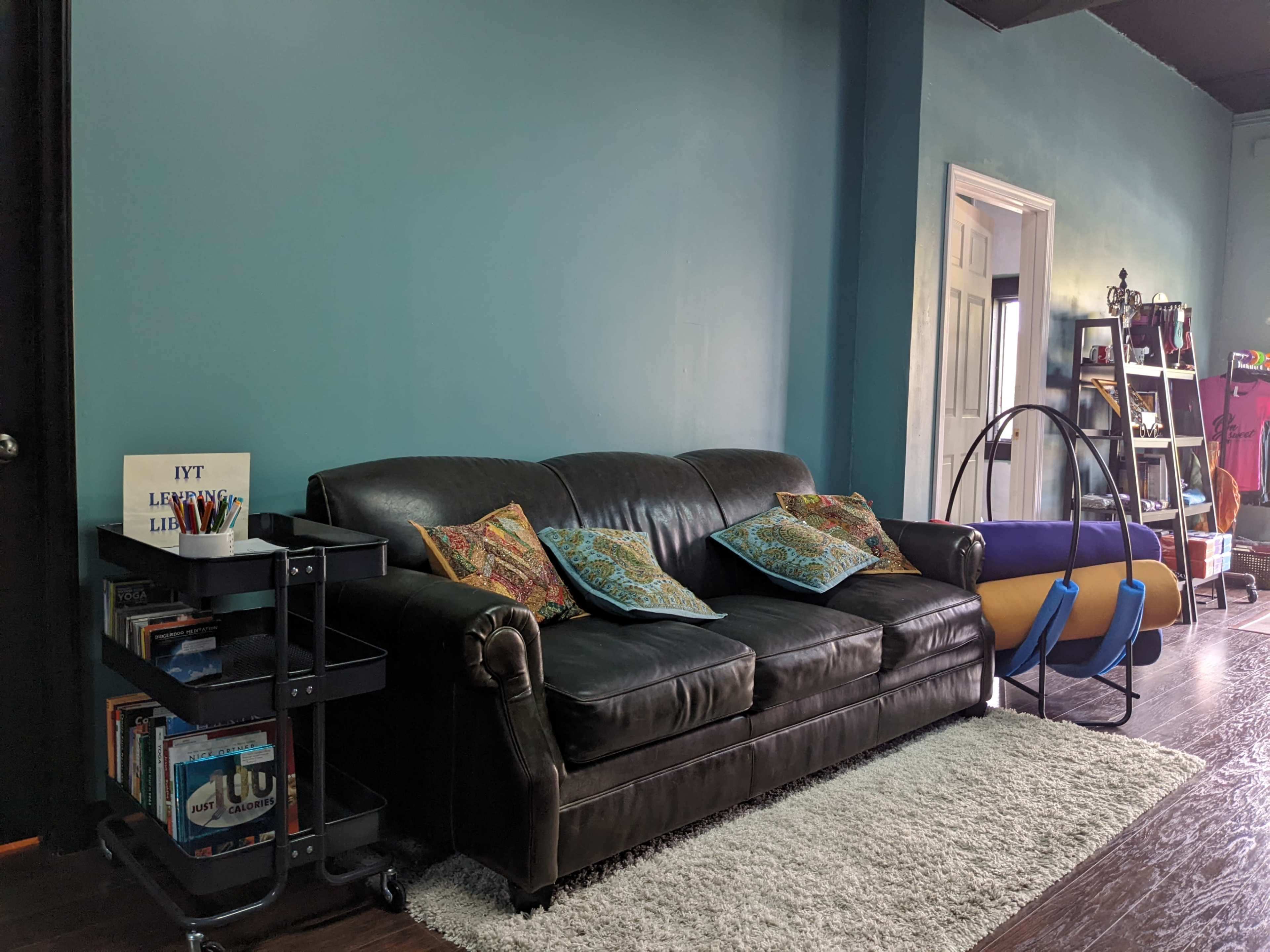 A living space featuring a dark leather couch with decorative pillows, a small black cart filled with books, a rug, and a yellow and blue structure next to a wall painted in teal.