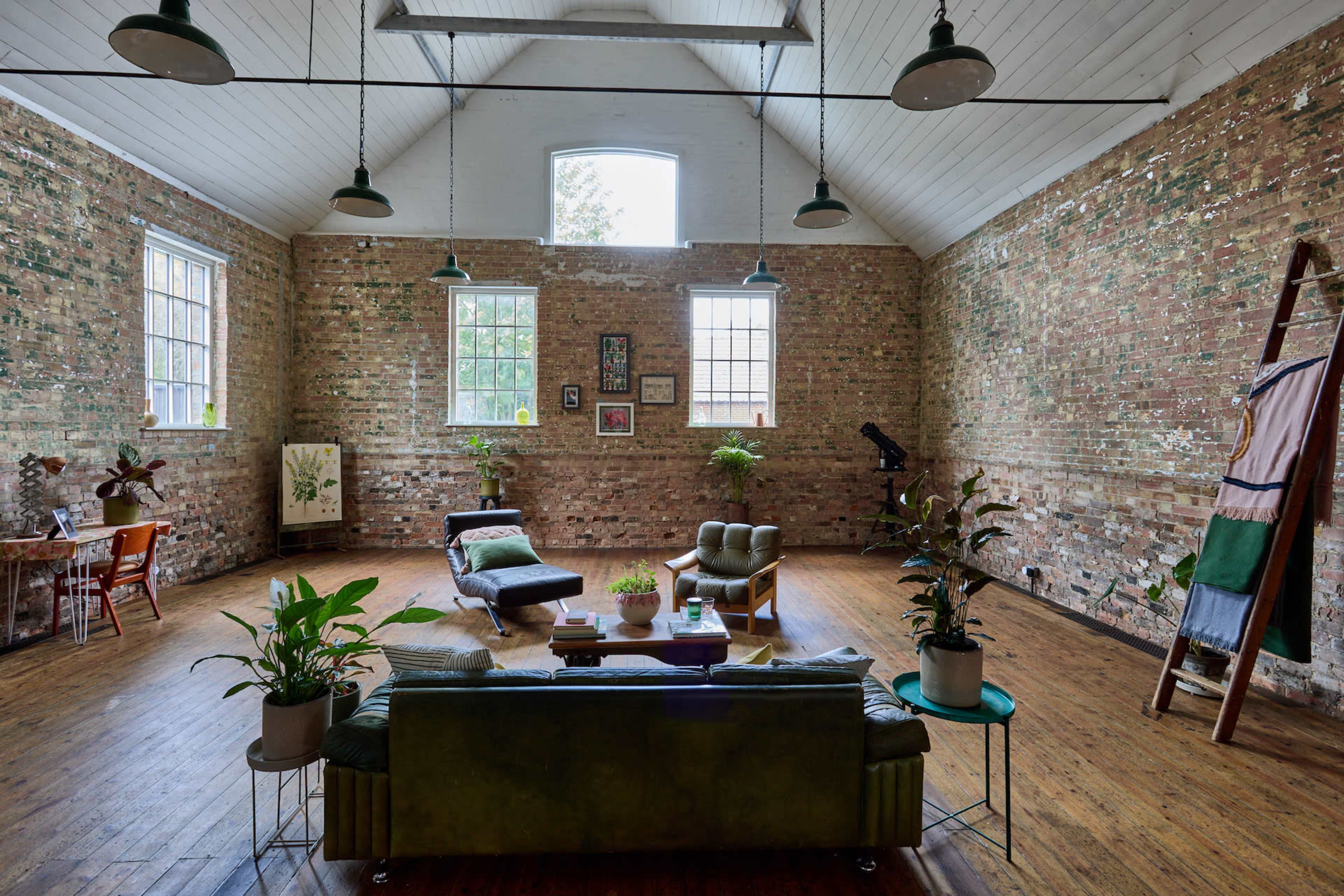 A spacious, open living area with exposed brick walls, large windows, and various plants arranged among minimalist furniture.