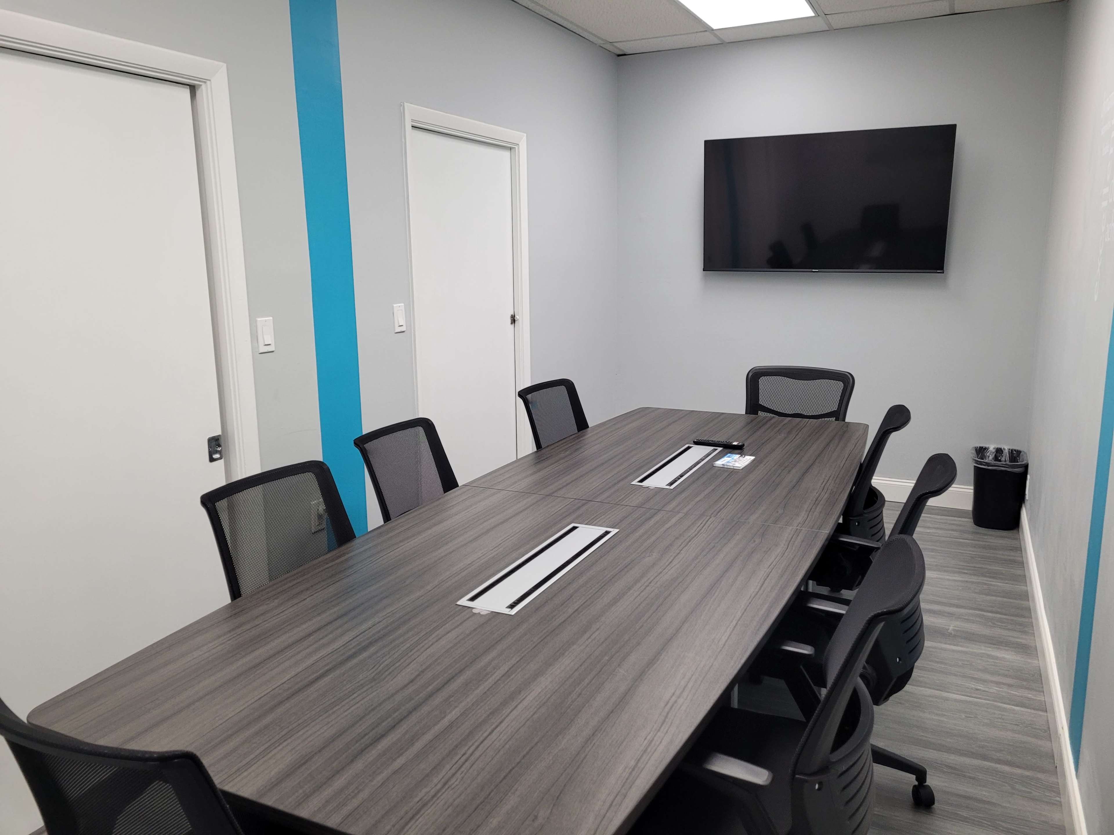 A modern conference room with a long table, several black mesh chairs, a wall-mounted television, and two doors.
