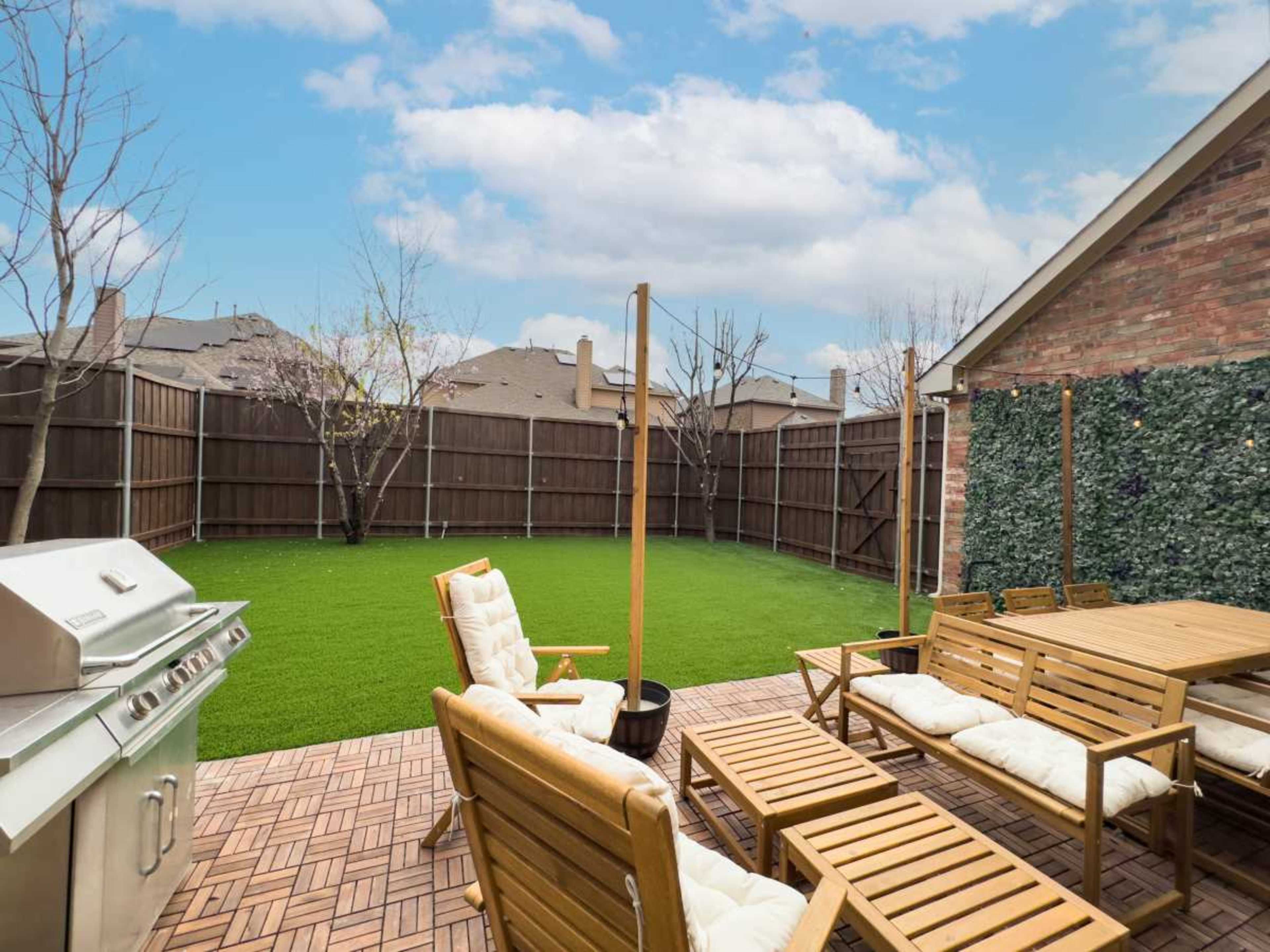 The image shows a backyard with a grill, wooden seating, and artificial grass, enclosed by a wooden fence.