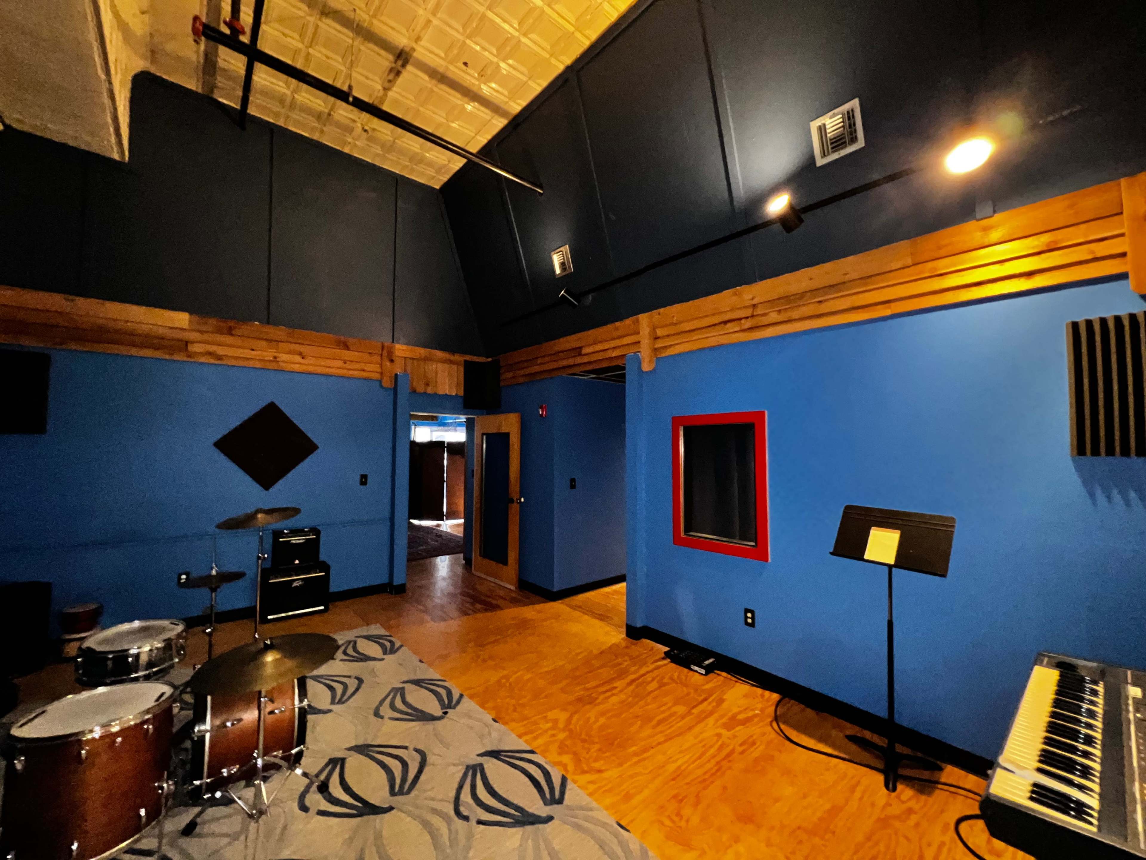 The image shows a recording studio with blue walls, wooden accents, and musical instruments including a drum set and keyboard.