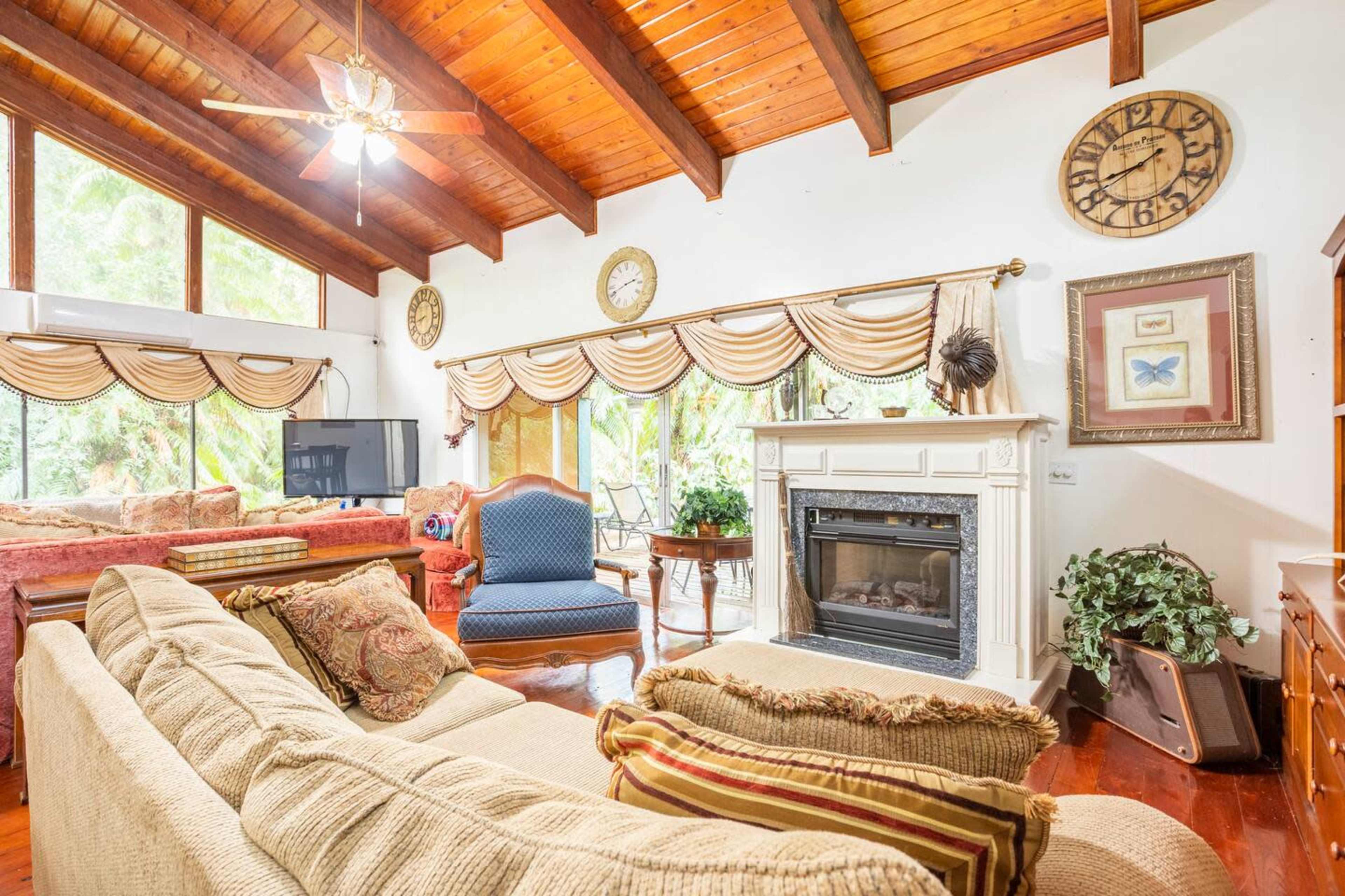 A living room features a beige sofa, a blue chair, and a fireplace beneath a wooden beam ceiling, with large windows allowing natural light to enter.
