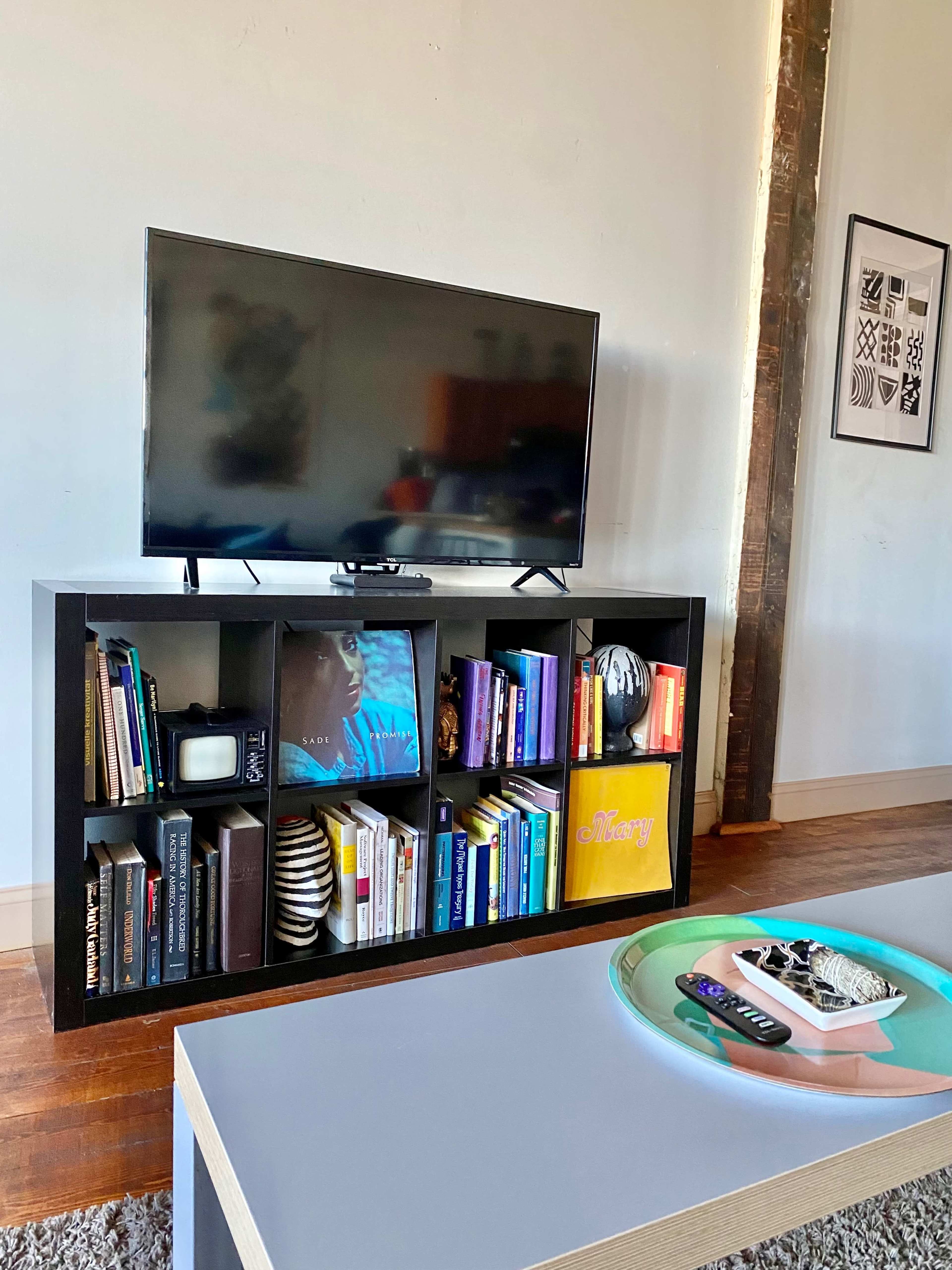 A television sits on a black shelf filled with books and decorative items in a well-lit living room.