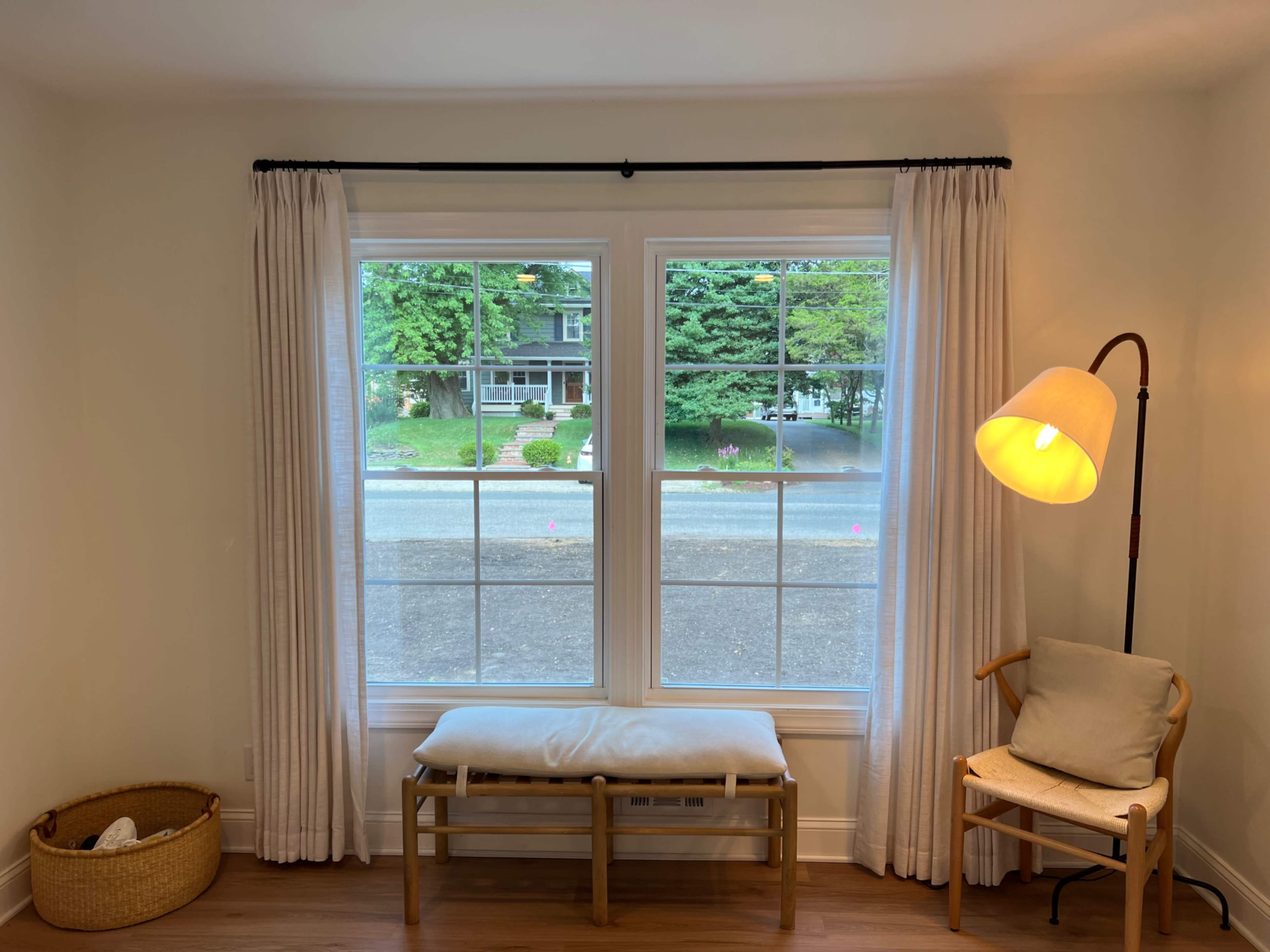 A cozy interior space with two large windows, a bench, a chair, a standing lamp, and a woven basket placed on the floor.