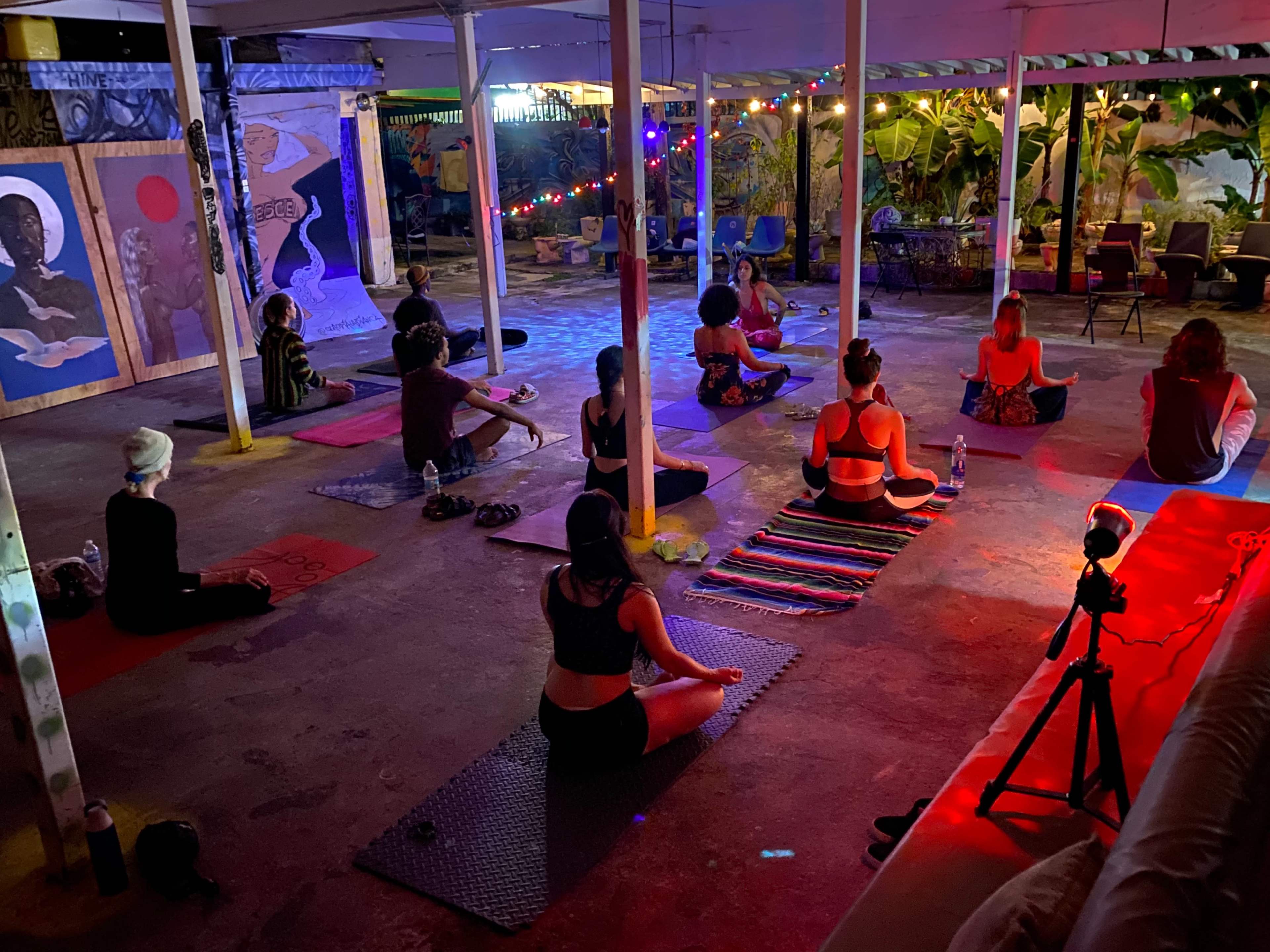 A group of individuals practices yoga in a dimly lit outdoor space with decorative lights and artwork in the background.