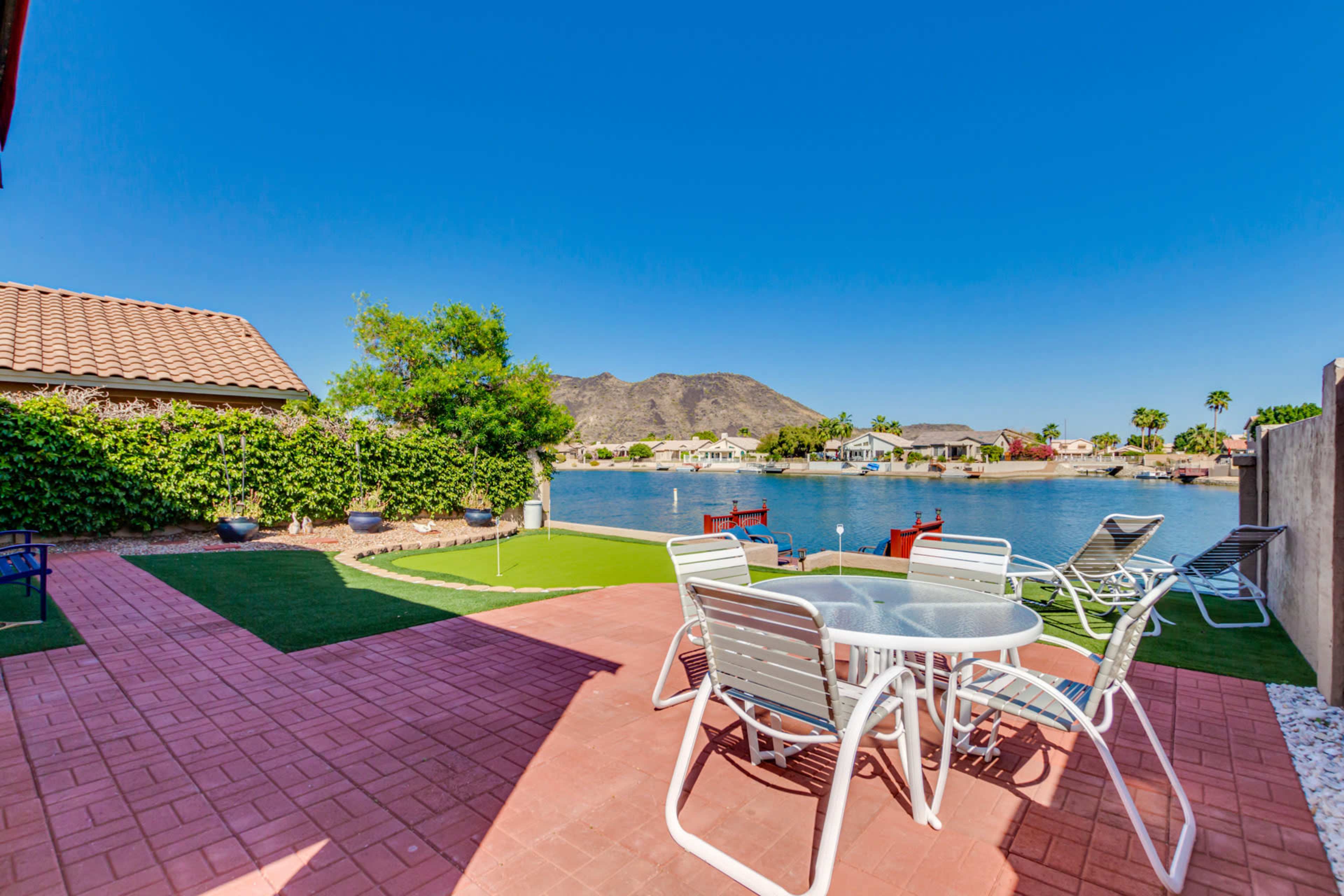 A patio with white metal chairs and a glass table overlooks a serene lake surrounded by houses and mountains under a clear blue sky.