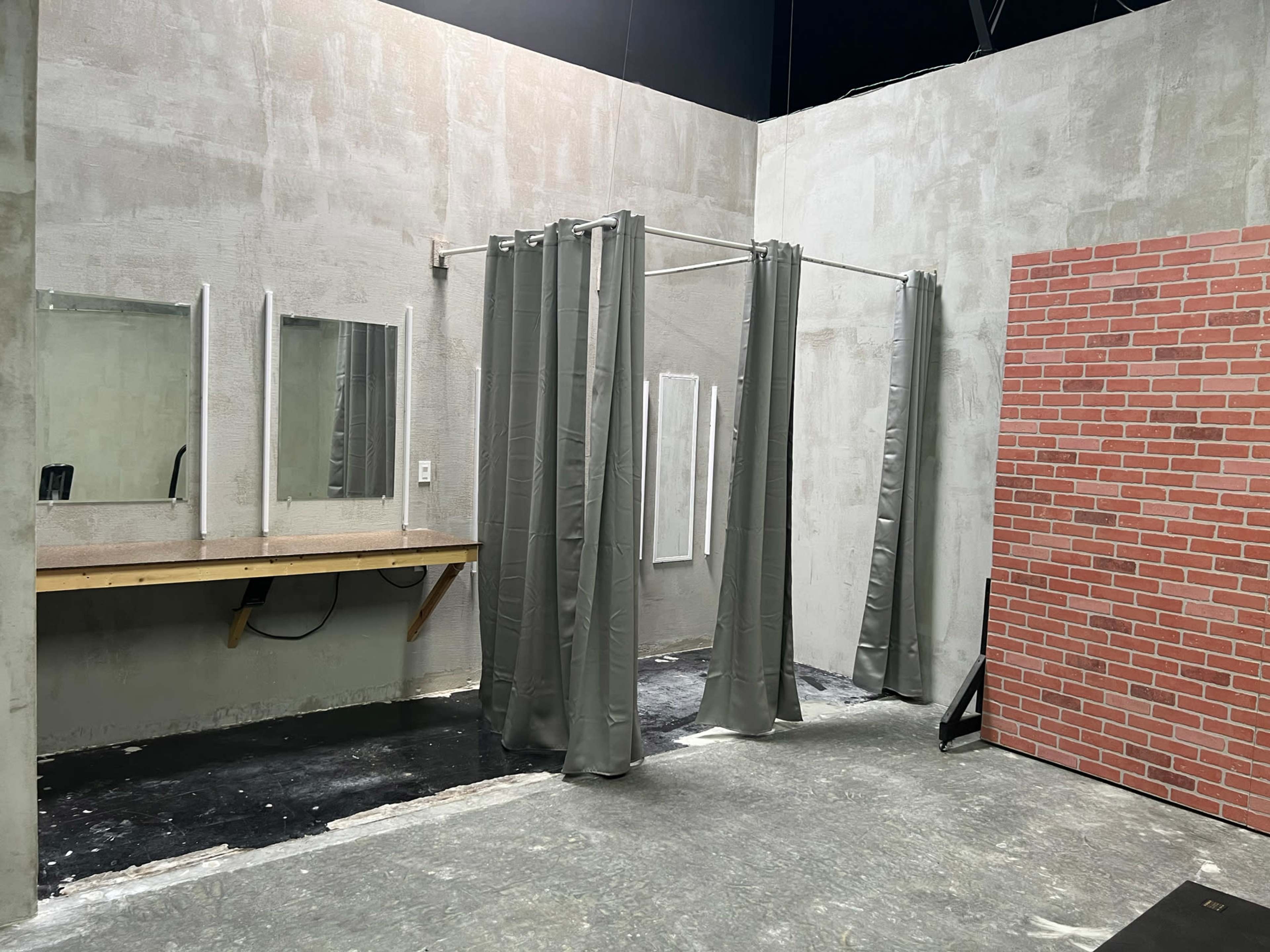 A studio space with gray walls, a wooden table, three mirrors, and shower curtains dividing areas, alongside a brick wall.
