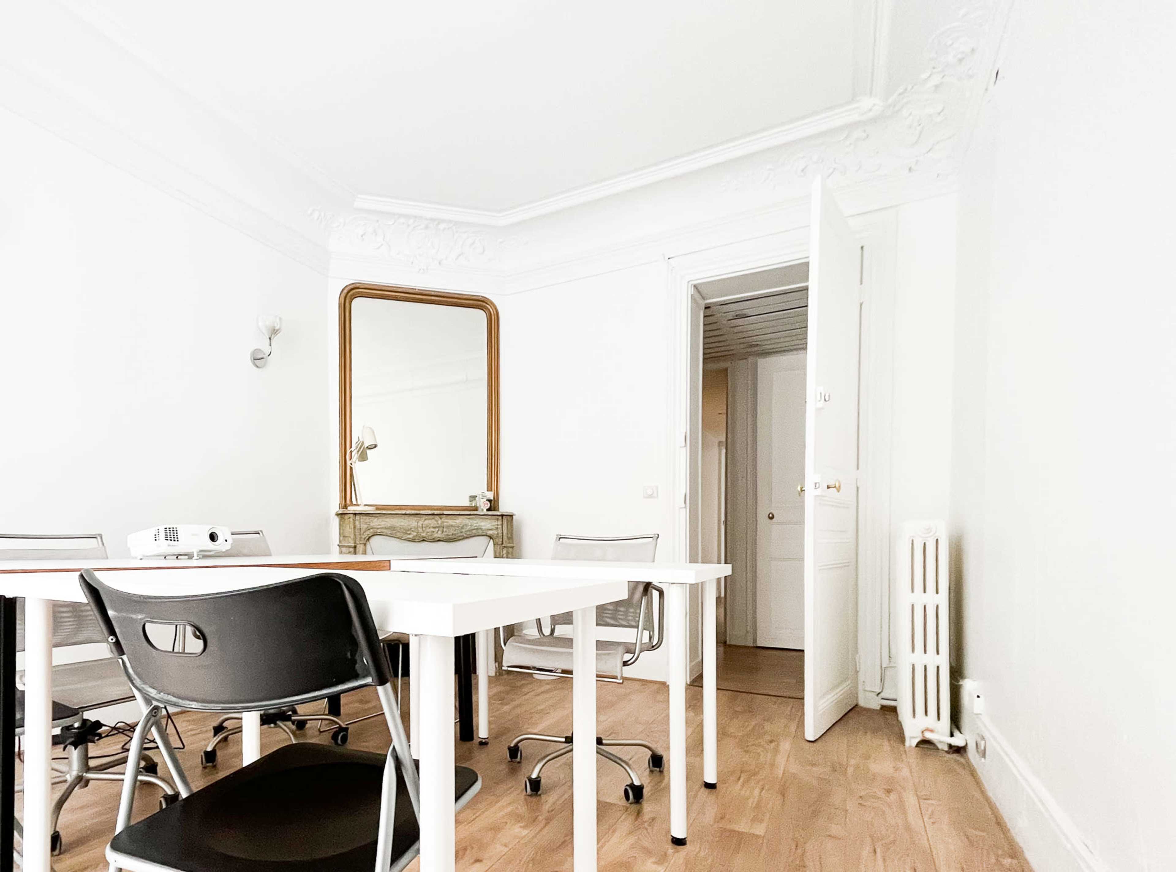 A minimalist office space featuring a white table, several black chairs, a large mirror, and an open door leading to another room.