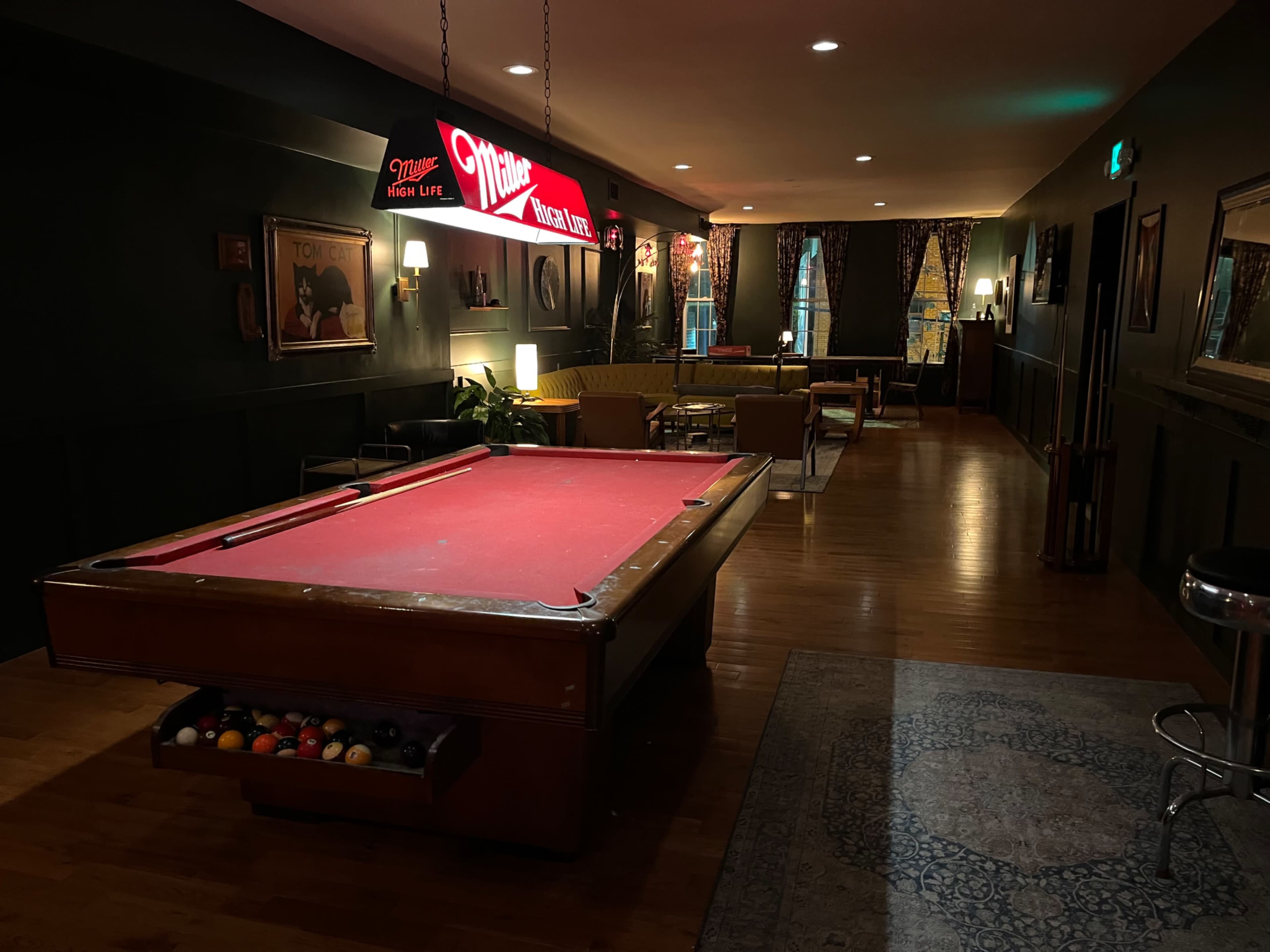 The image shows a dimly lit room featuring a red pool table, a bright pool hall sign above it, and a seating area with yellow couches and curtains in the background.