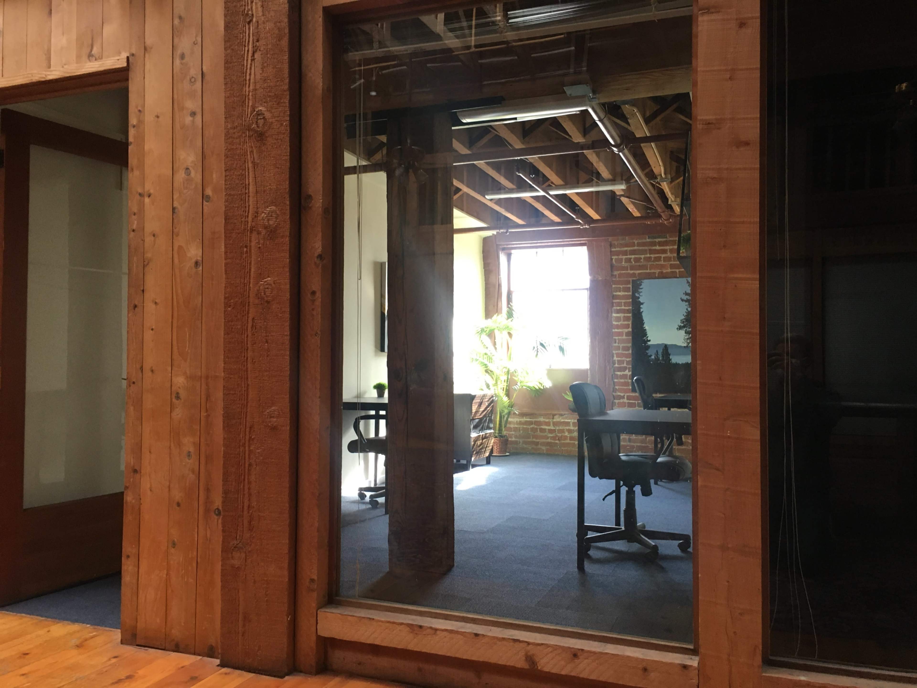 The image shows a wooden office space with glass walls, featuring two chairs and a large window allowing natural light to enter.