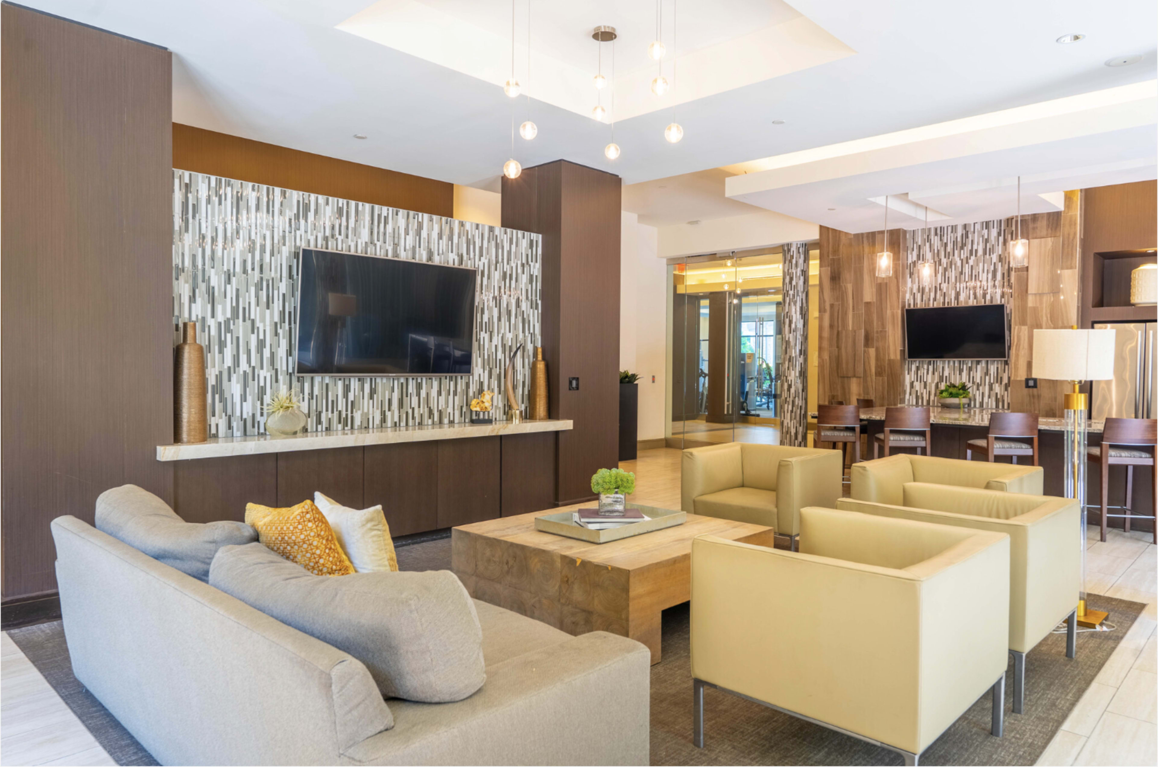 The image shows a modern lobby area with a gray sofa, beige armchairs, a wooden coffee table, and a television mounted on the wall, accented by decorative lighting.