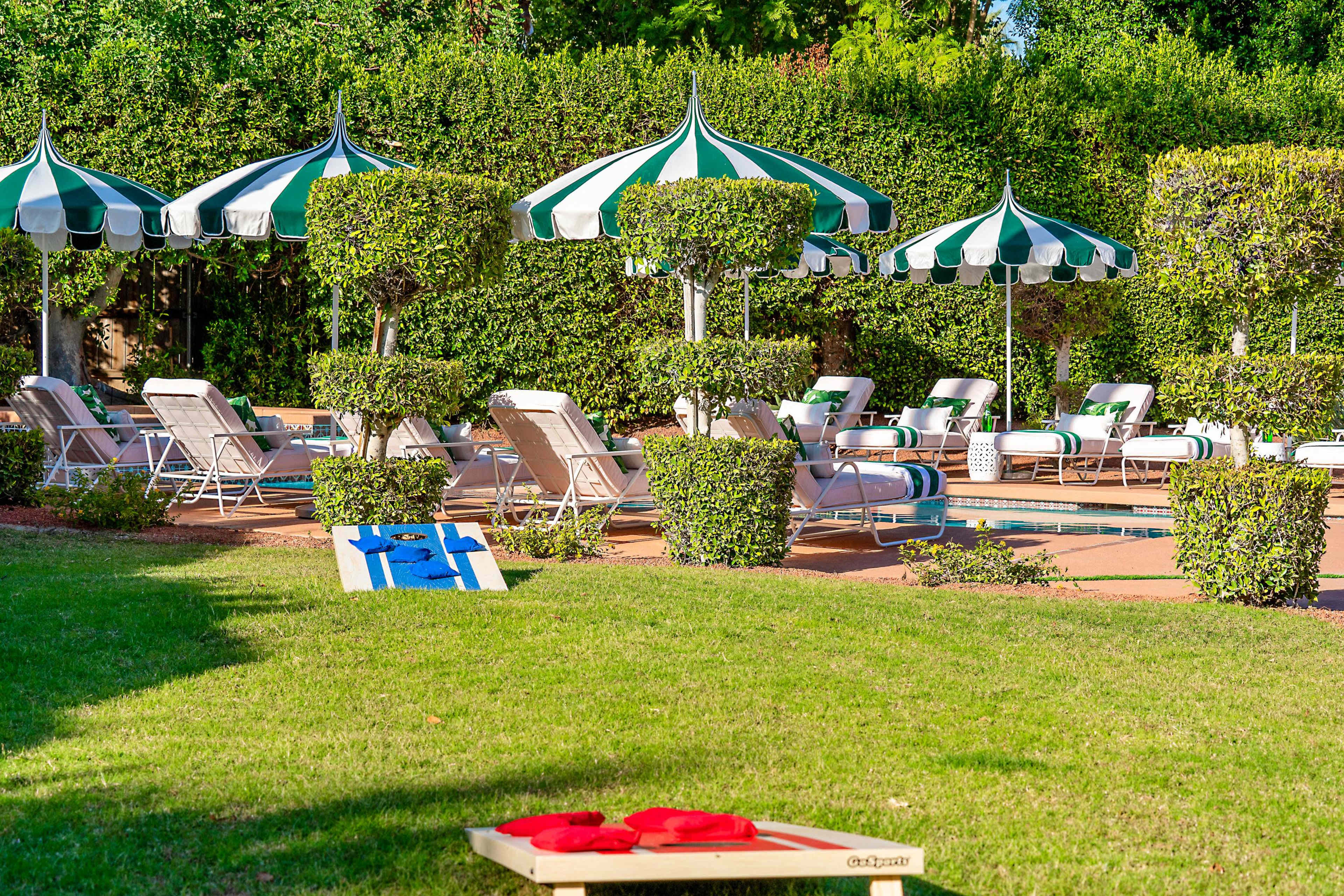 The image shows a poolside area with striped umbrellas, lounge chairs, and a cornhole game set up on the grass.
