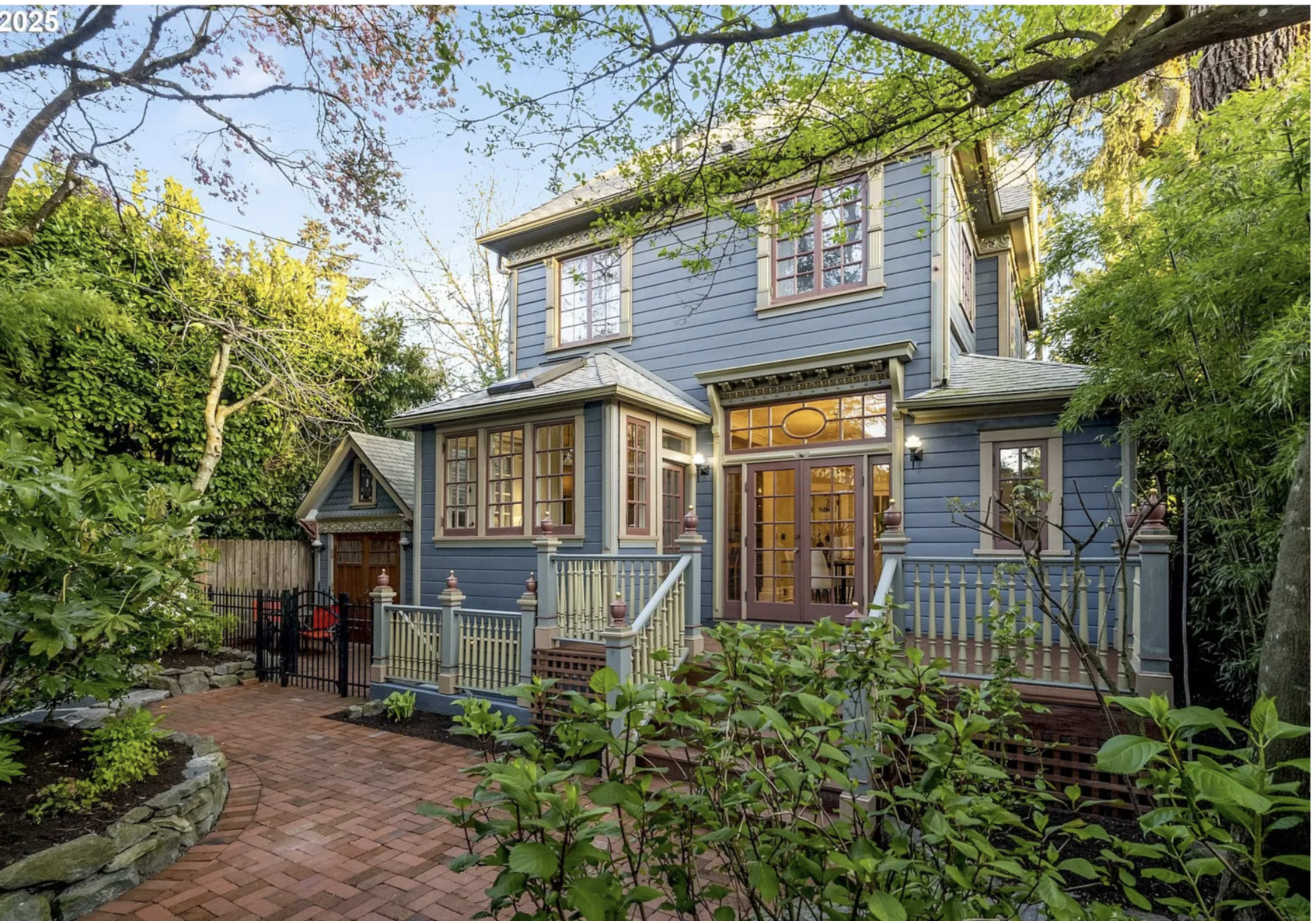 A two-story blue house with multiple windows and a front porch is surrounded by lush greenery and a brick walkway.