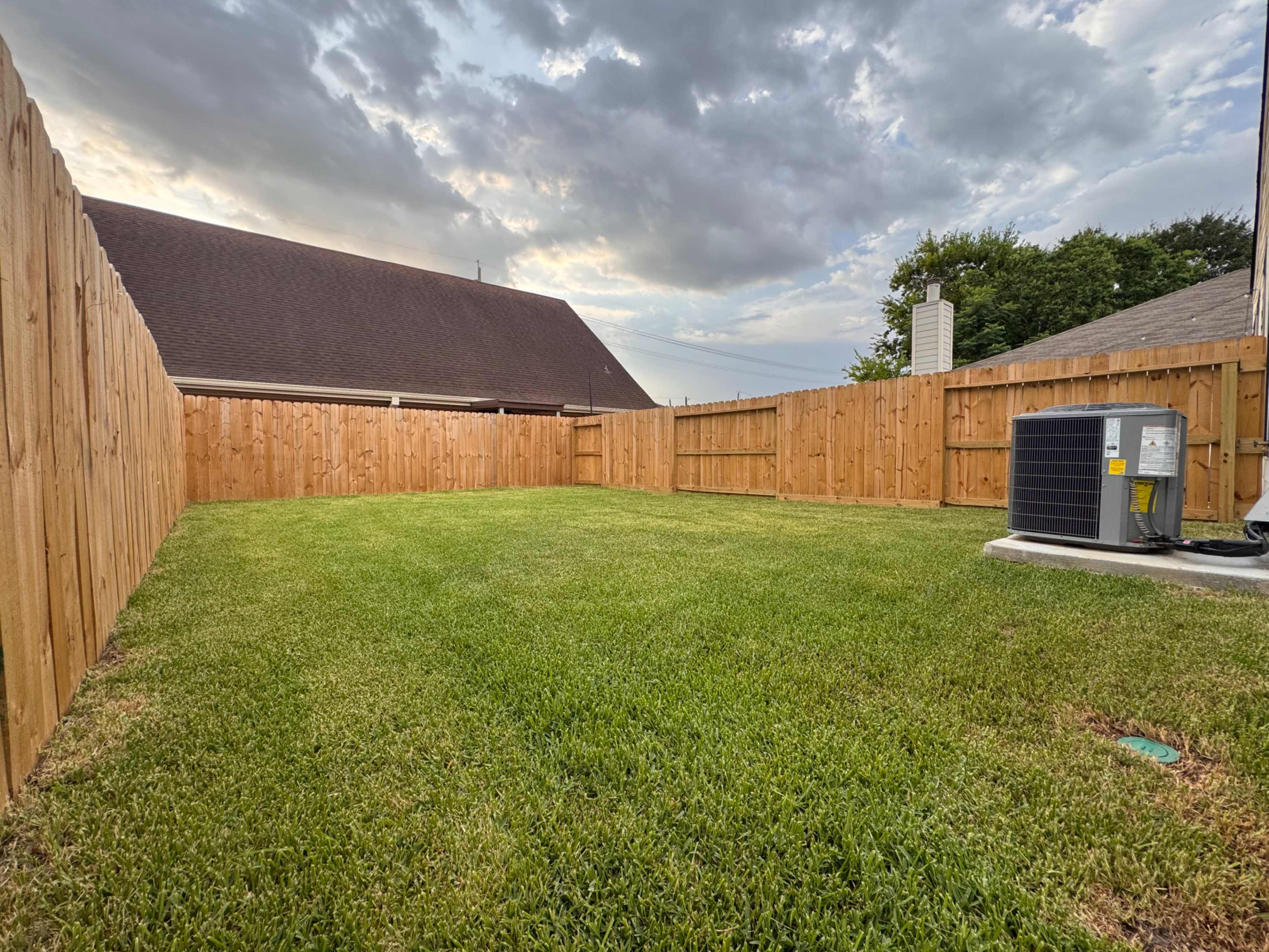 The image shows a fenced backyard with a neatly manicured lawn and an air conditioning unit in one corner.