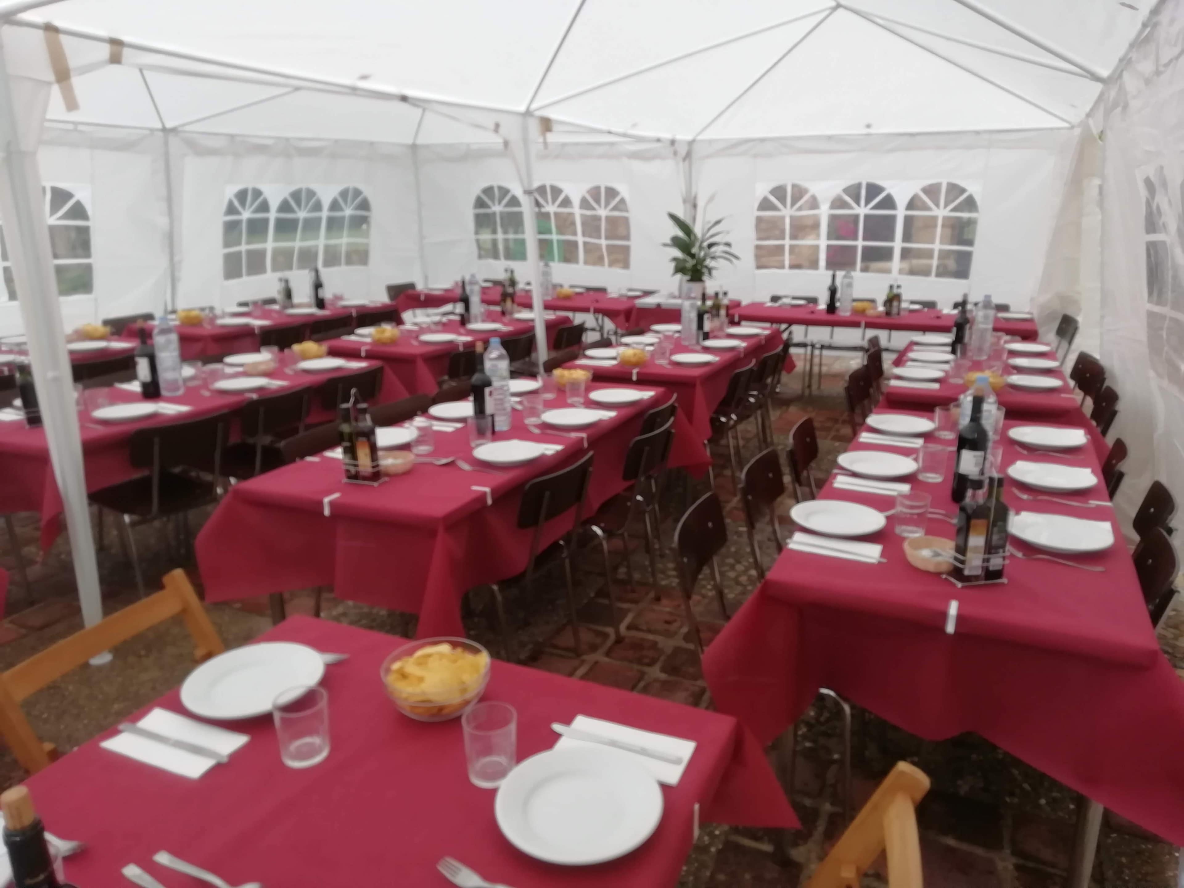 A large tent is set up with multiple tables covered in red tablecloths, each arranged with plates, glasses, and bottles.