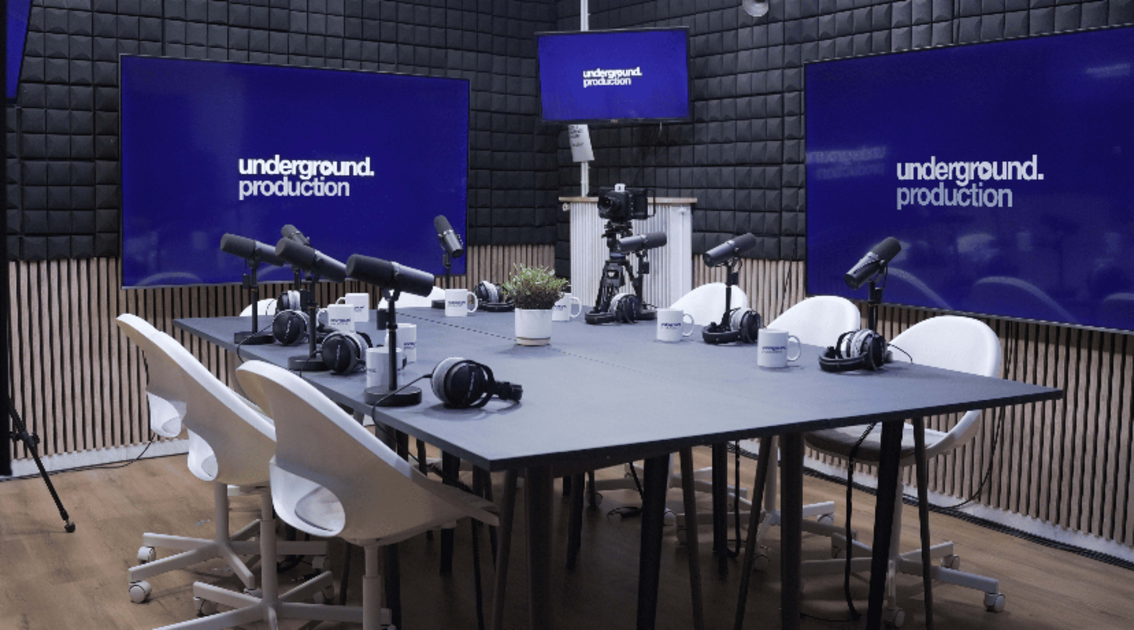 A modern podcast studio features a large table with microphones, headphones, and mugs, surrounded by multiple screens displaying the text "underground.production."