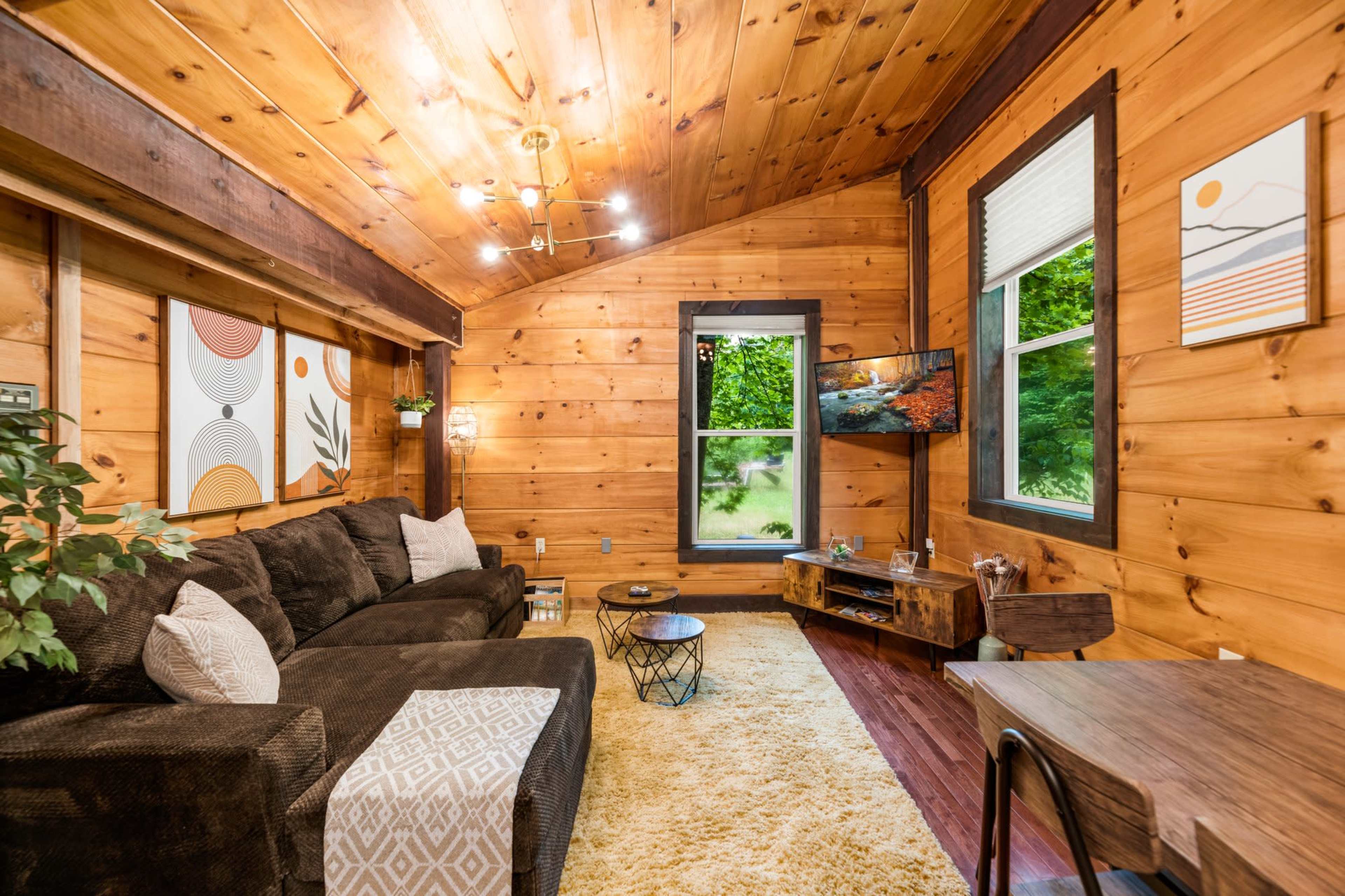 A cozy wooden room featuring a dark sectional sofa, a coffee table, and a television mounted on the wall, with large windows allowing natural light to enter.