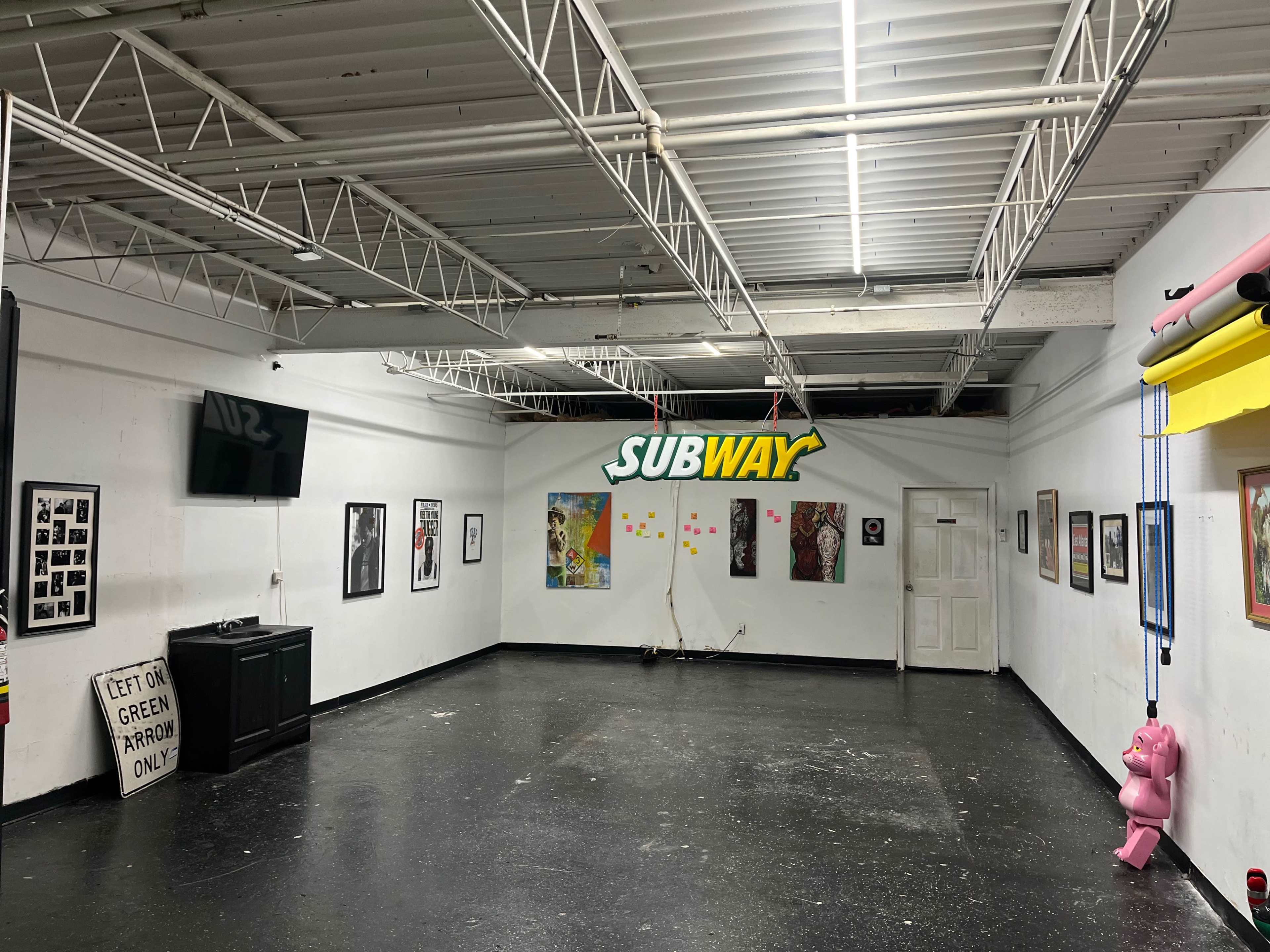 The image shows a modern art gallery with a black floor, white walls adorned with various framed artworks, and a prominent Subway sign hanging from the ceiling.