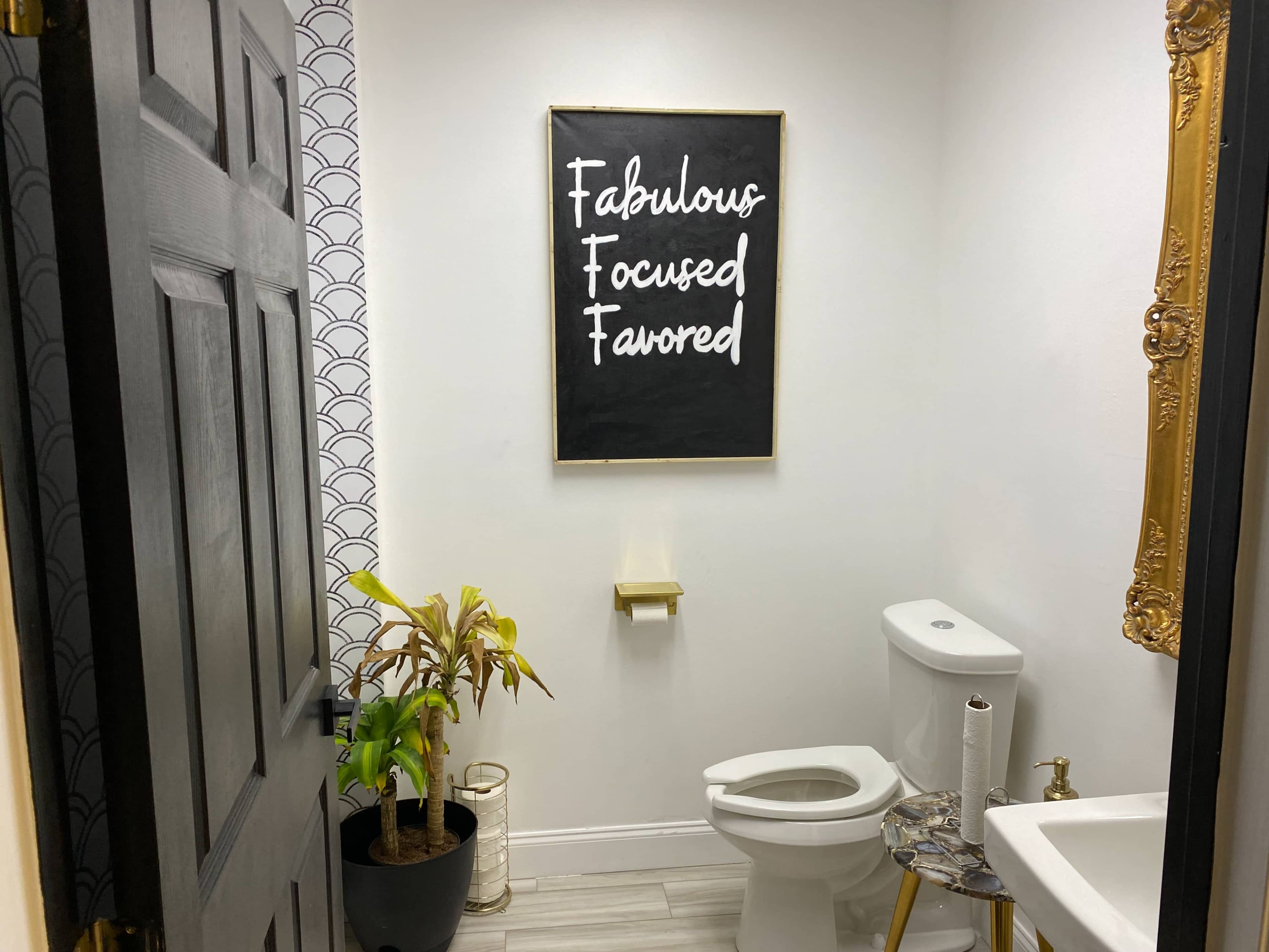 A small bathroom features a blackboard sign with inspirational words, a toilet, and a potted plant beside the door.