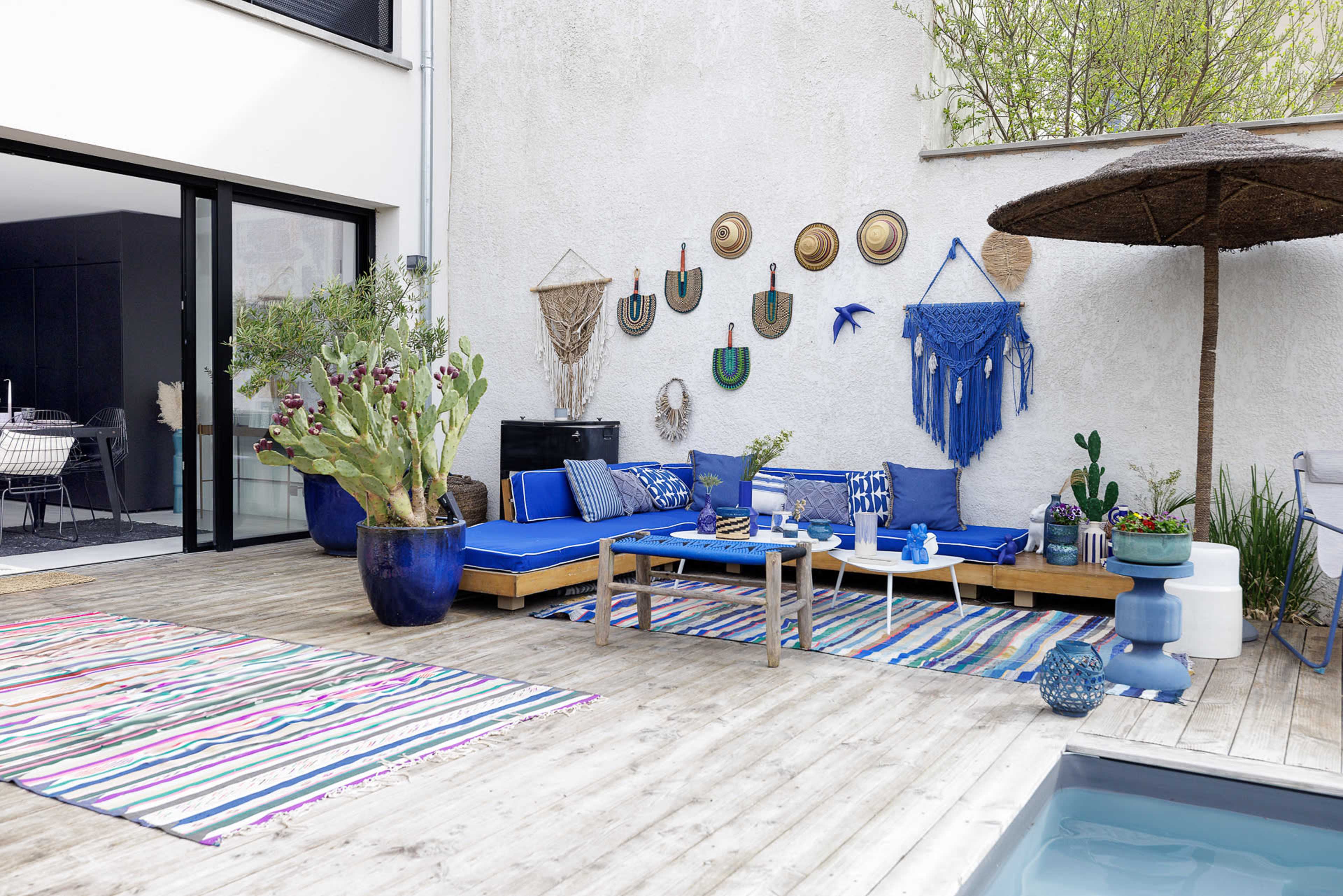 A stylish outdoor patio equipped with a wooden deck, a blue seating area, decorative wall hangings, and a small pool.