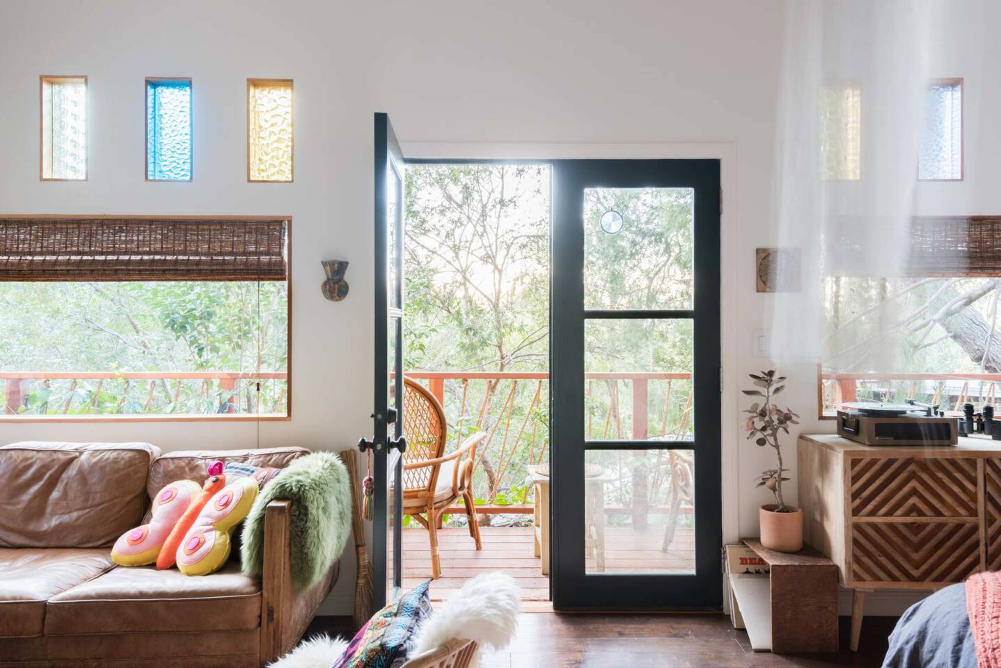 A cozy room features a brown leather sofa, colorful cushions, and a door opening to a wooden balcony surrounded by trees.