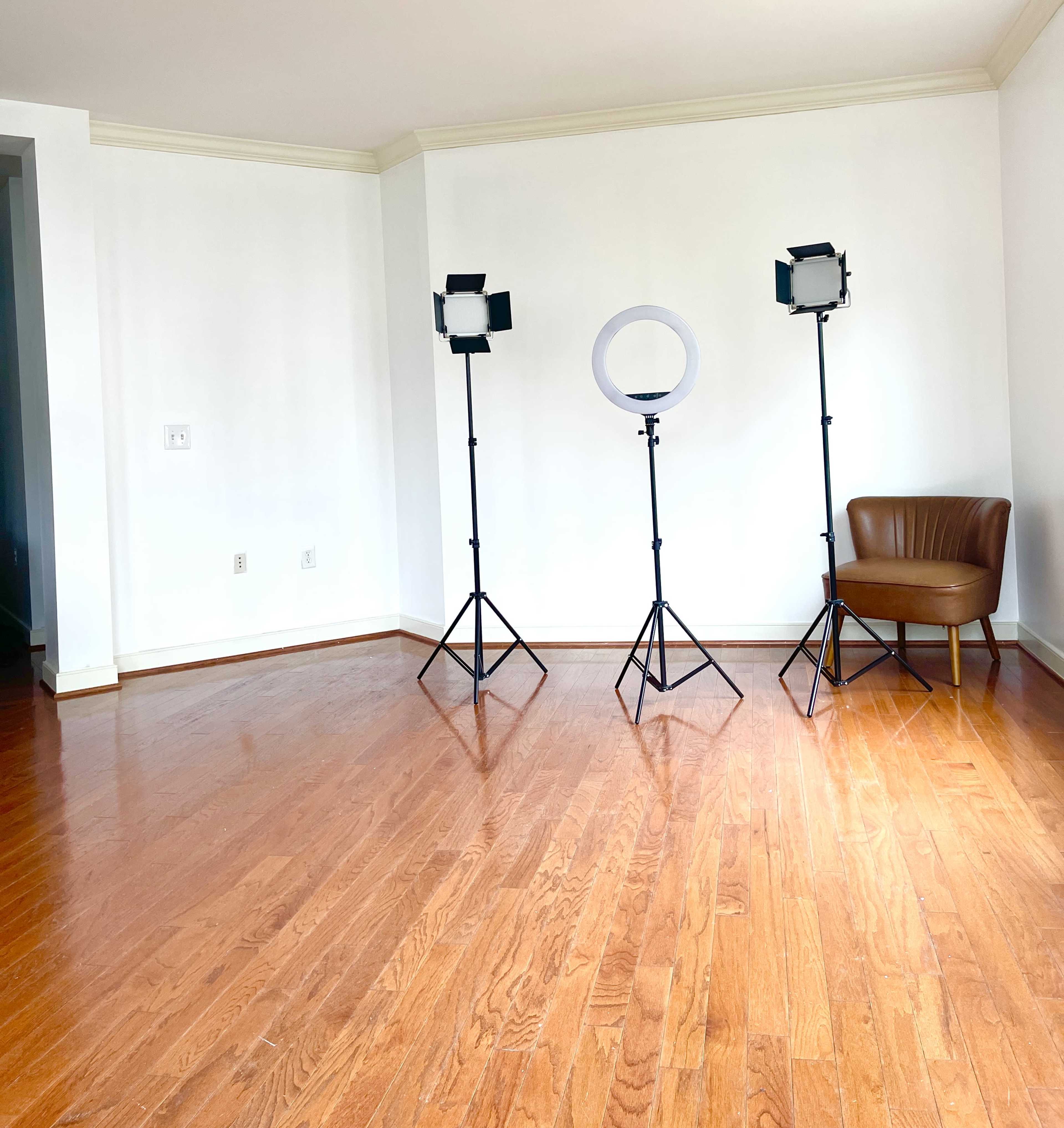 The image shows a spacious, empty room with wooden flooring, featuring three lighting stands and a brown chair against a white wall.