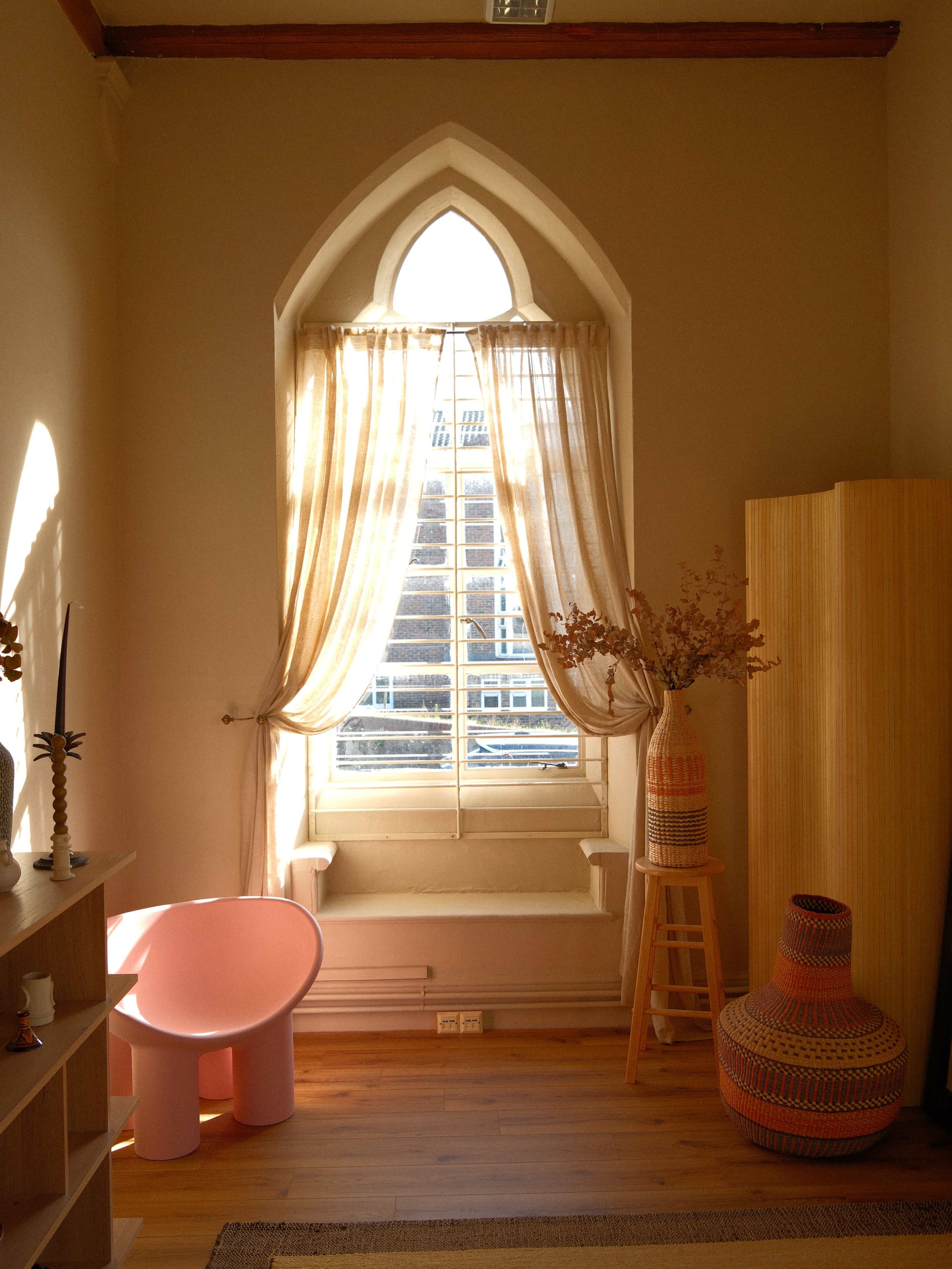 The image shows a bright room with an arched window dressed in light curtains, a pink chair, a wooden stool, and decorative vases arranged near the window.