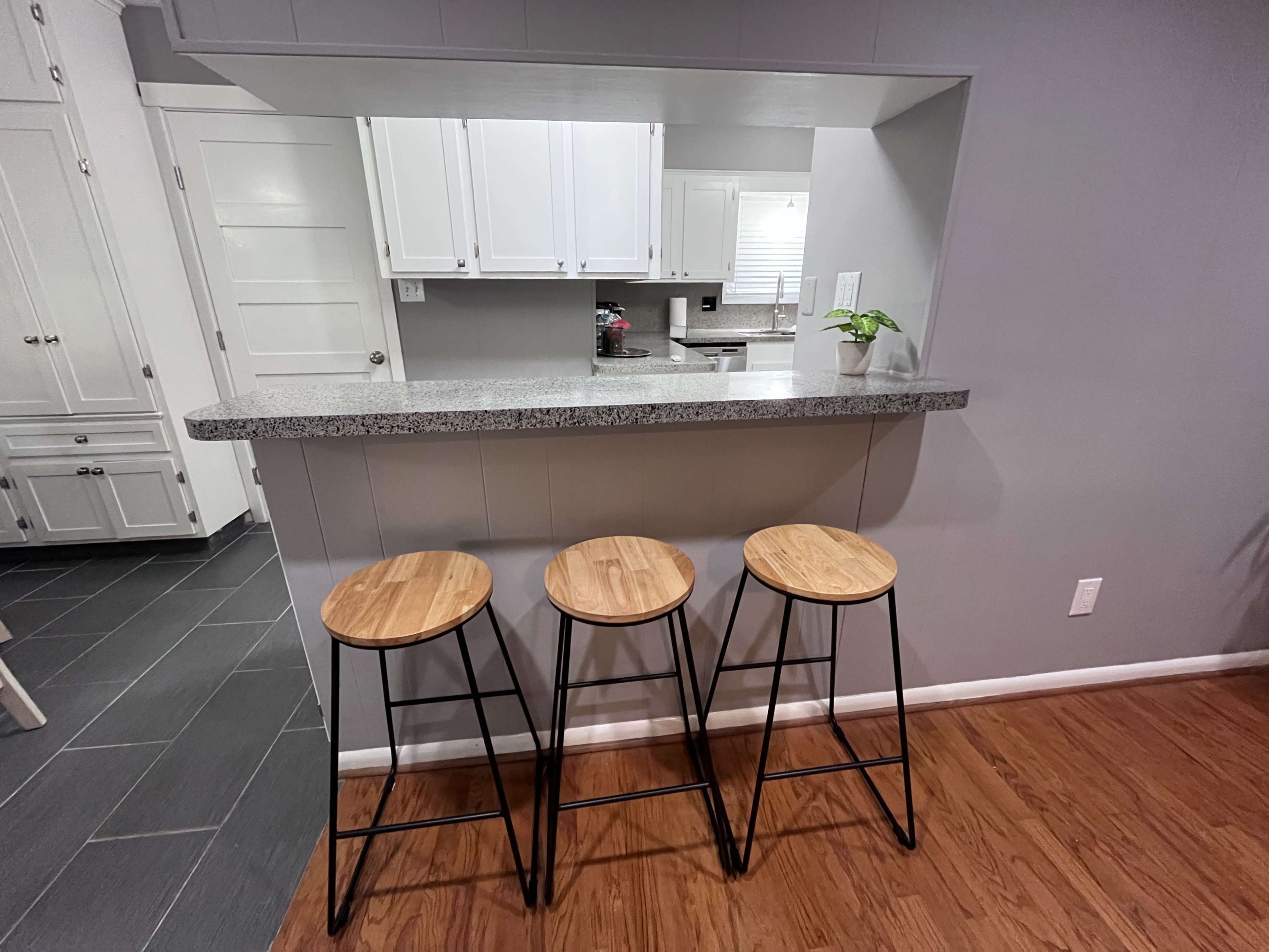 The image shows a kitchen counter with three wooden bar stools in front of it, set against a gray wall.