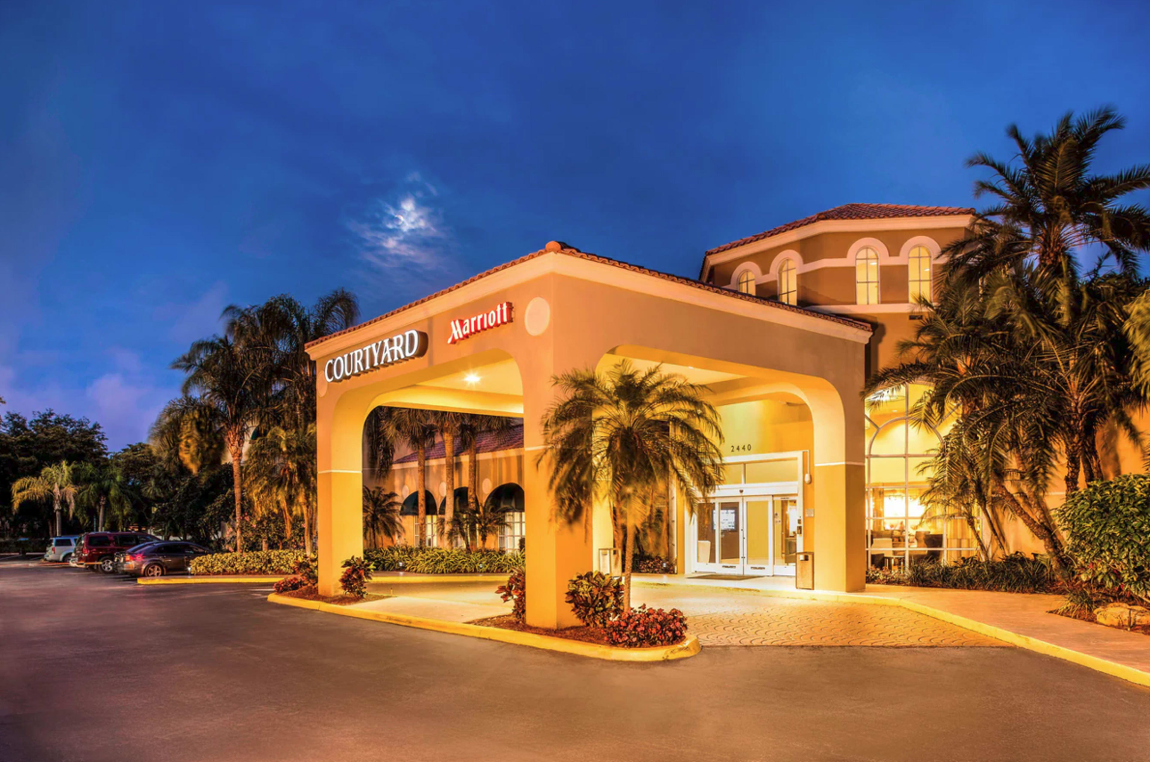 The image shows the entrance of a Courtyard by Marriott hotel, featuring a portico with palm trees and parking spaces in the foreground during twilight.