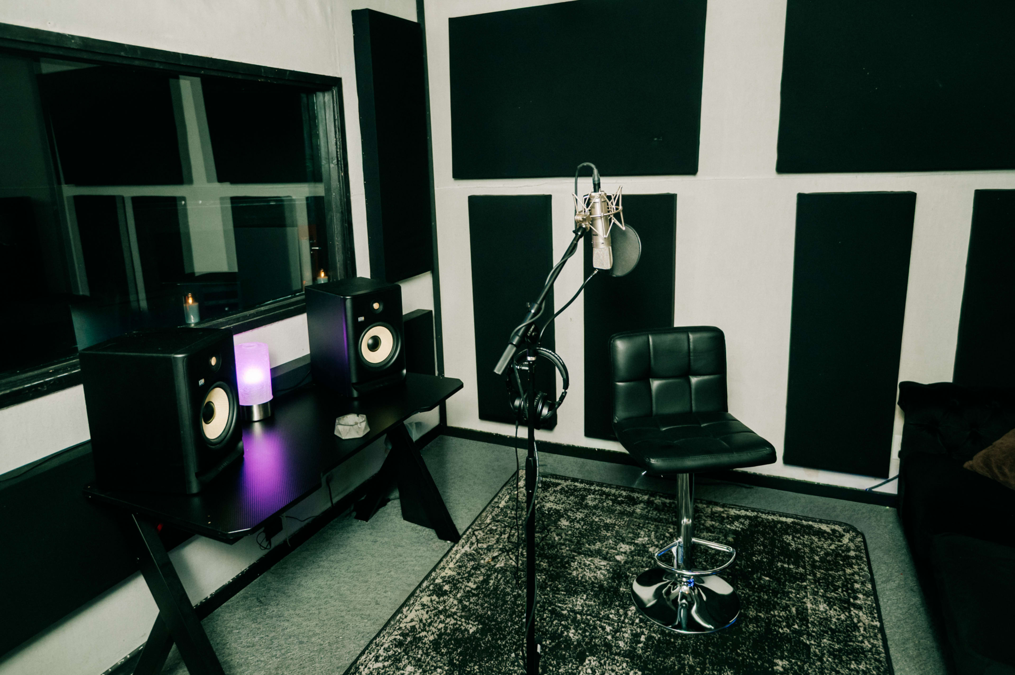 The image shows a music recording studio with a microphone on a stand, a black chair, and audio monitors on a table.