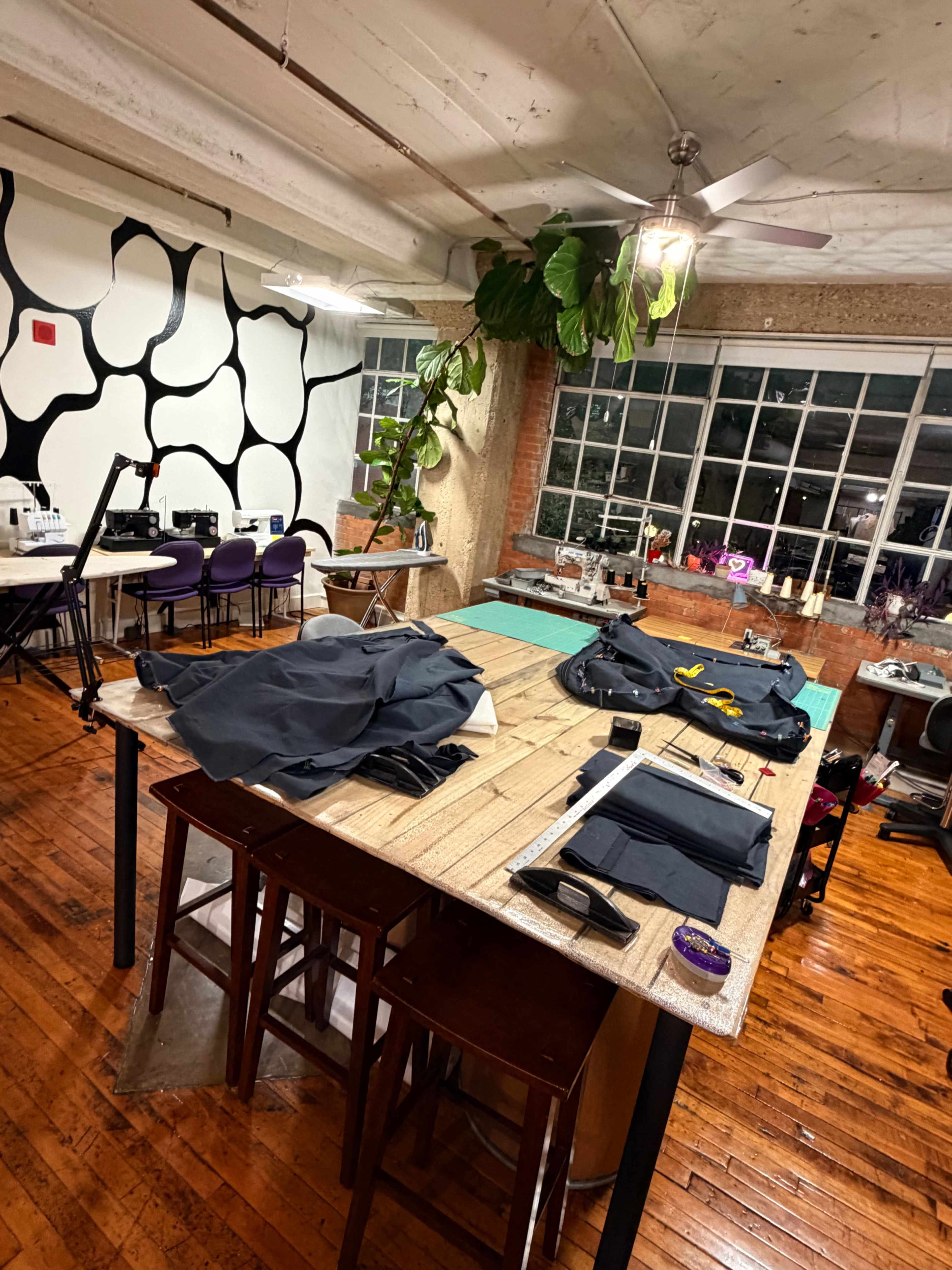 The image shows a spacious sewing studio with a large wooden table covered in fabric and tools, surrounded by machines and a vibrant wall design.