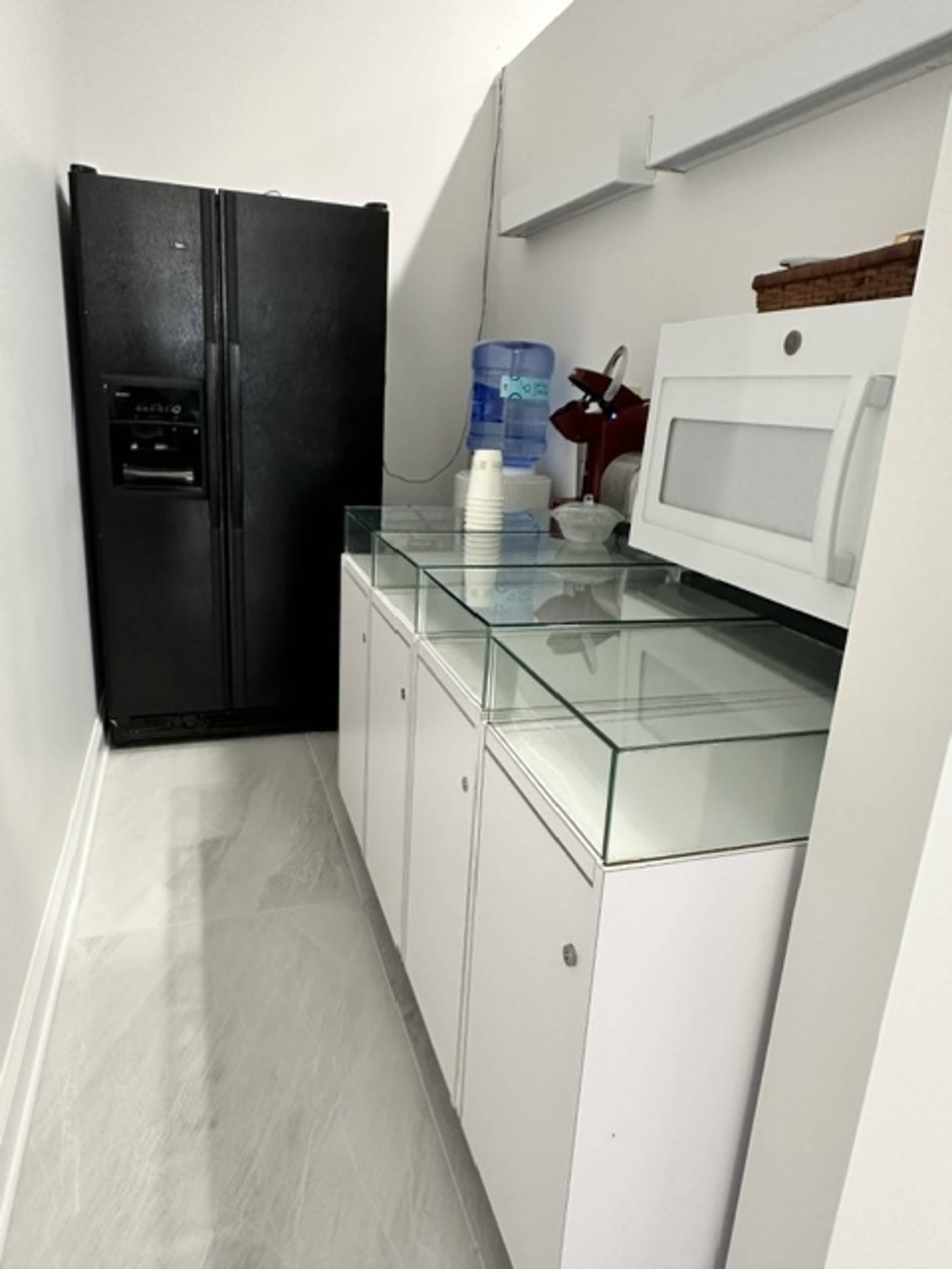 The image shows a kitchen corner featuring a black refrigerator, a glass-topped cabinet, a microwave, and a water dispenser.